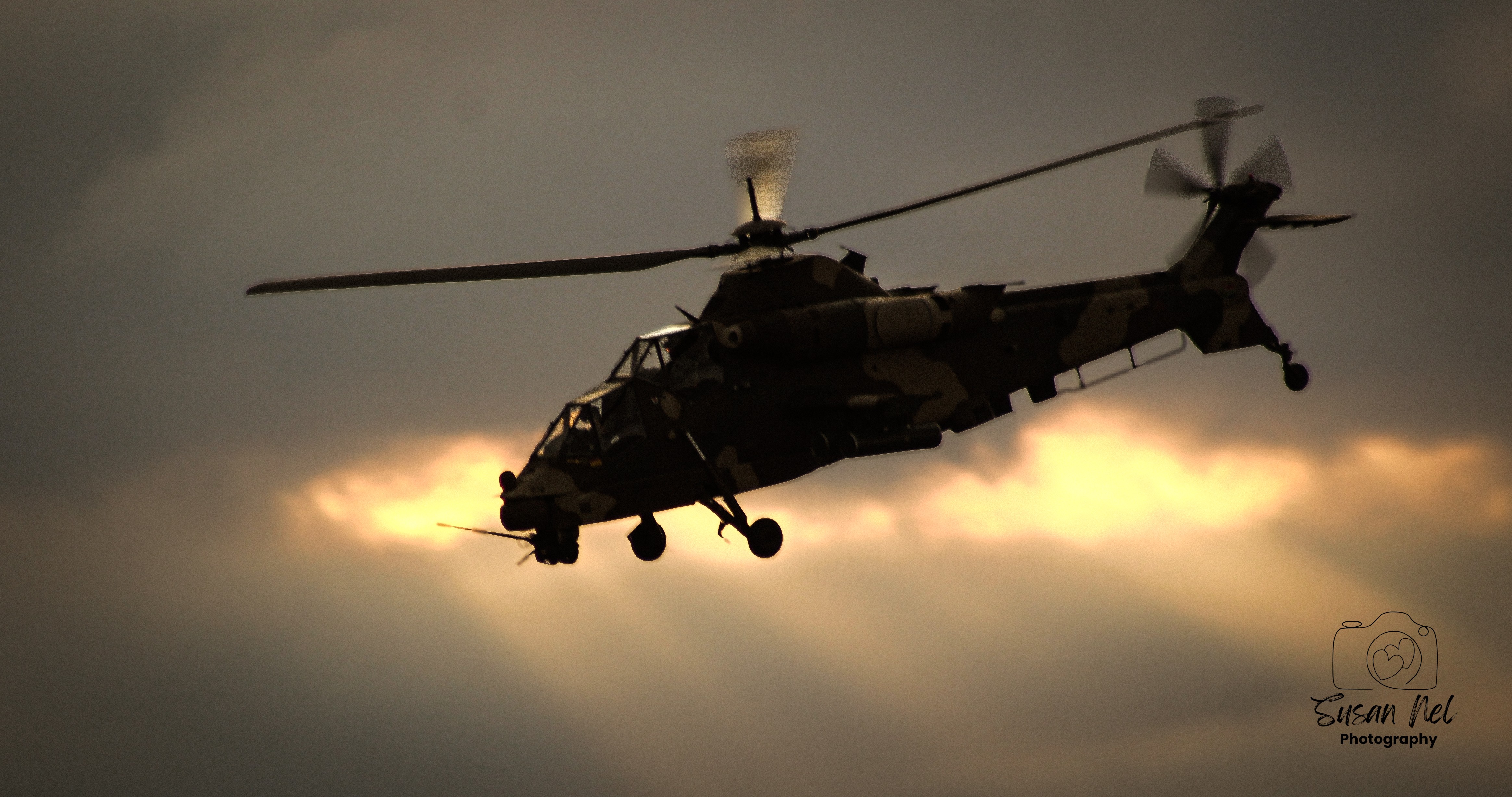 A South African  Airforce Rooivalk military attack helicopter of 16 Squadron flying through the air, silhouetted against a dramatic, cloudy sky. The combination  of the dark aircraft, glowing clouds and blurred rotor blades captures the power and atmosphere of the Rooivalk in flight during the Africa Aerospace and Defence 2026 exhibtion at AFB Waterkloof, South Africa.