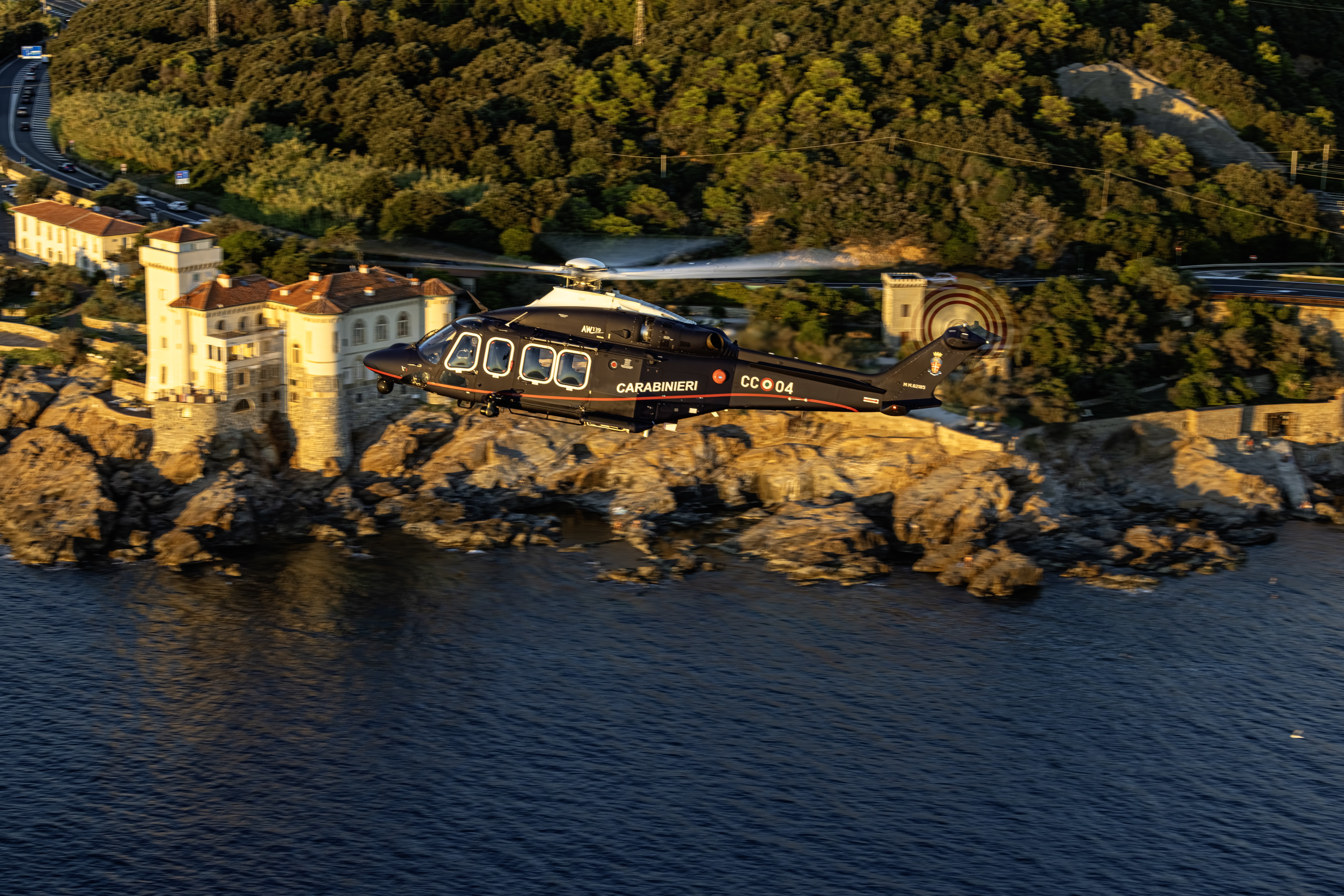 A Carabinieri (military police) UH-149D helicopter during the "golden hour" along the Livorno coast, monitoring road and marine traffic amidst the area's distinctive rocky coastline.
