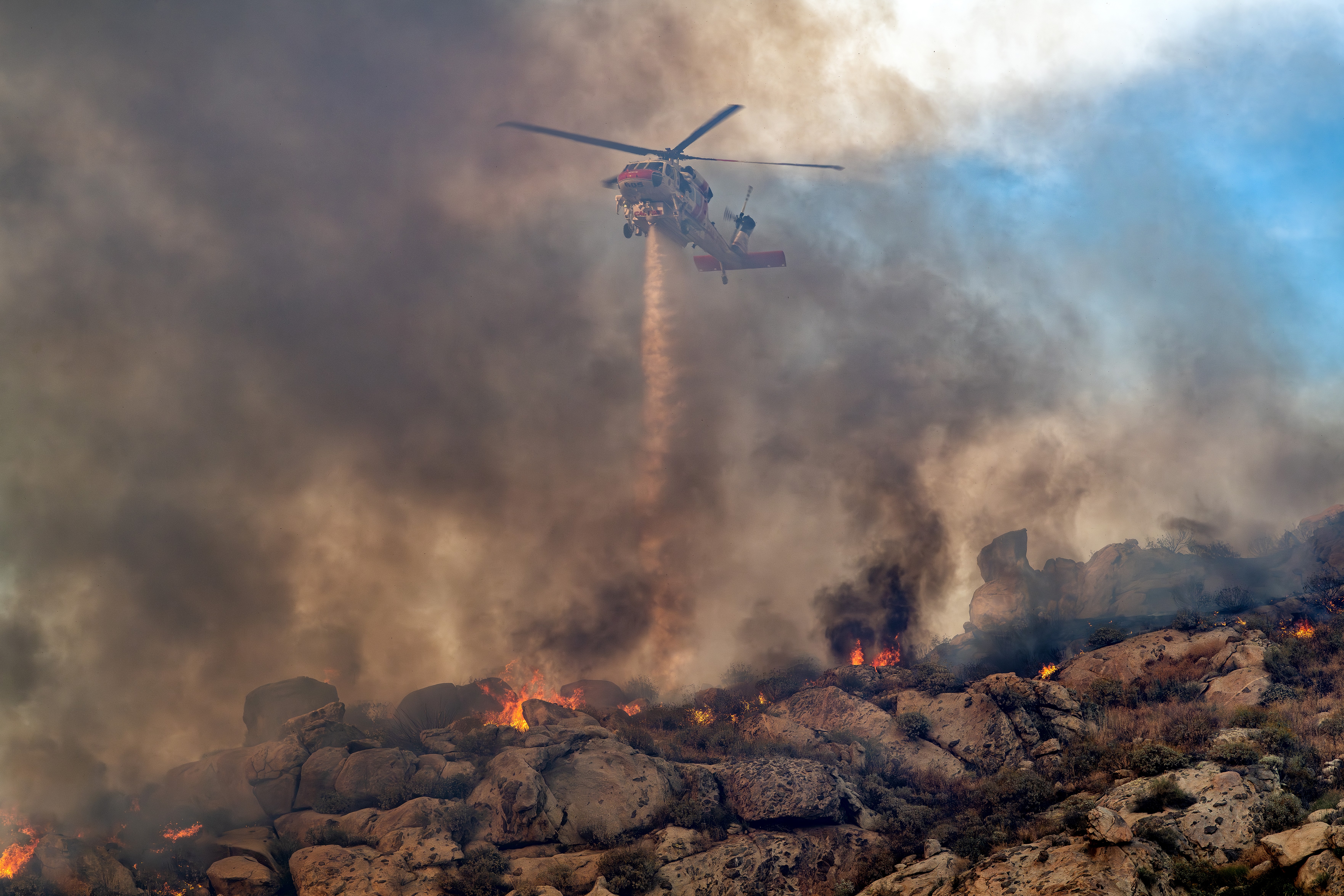 It is difficult for my camera to focus when an aircraft is behind a smoke-filled sky, so I prefocused the camera, turn off my auto focus, and waited for the right composition. The smoke-filled sky and fire burning below made for a really dramatic photograph of Cal Fire's Firehawk dropping water. Taken at Lake Perris, California during the Springs Fire. The area we had to drive around to get aircraft dropping was pretty wide and the app "Watch Duty" was used to get us to the right location. To get to the best position for photographing, we had to drive on a dirt road making sure we were not in the way of the fire trucks. One of the most important things about photographing fires is situational awareness.