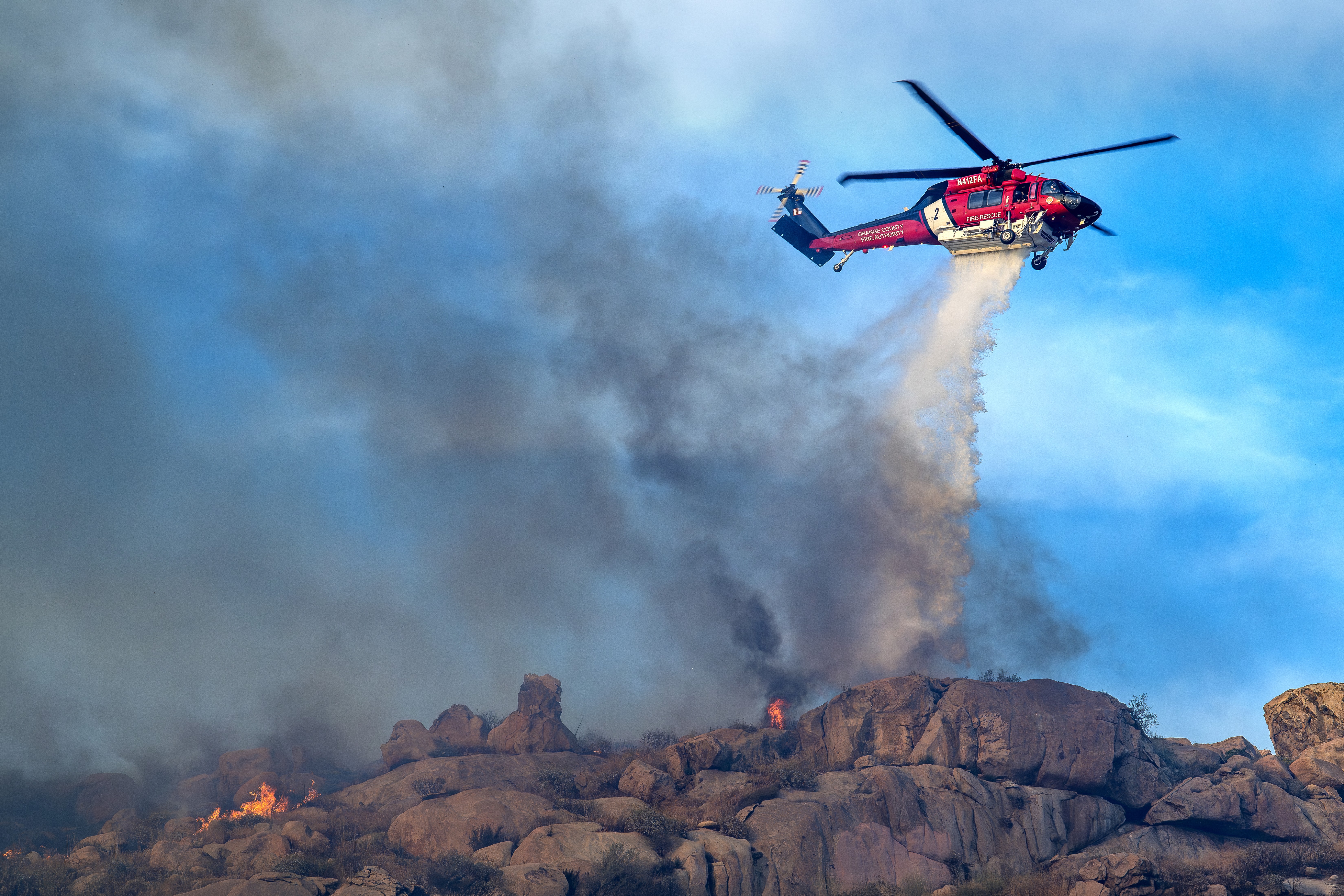 Orange County Fire Authority's Firehawk drops on the Springs Fire in Lake Perris, California. The late afternoon light was very soft and perfectly illuminated the Firehawk and falling water against a beautiful blue sky. I shot this photograph at a wider angle to include the smoke and burning landscape. The area we had to drive around to get aircraft dropping was pretty wide and the app "Watch Duty" was used to get us to the right location. To get to the best position for photographing, we had to drive on a dirt road making sure we were not in the way of the fire trucks. One of the most important things about photographing fires is situational awareness.