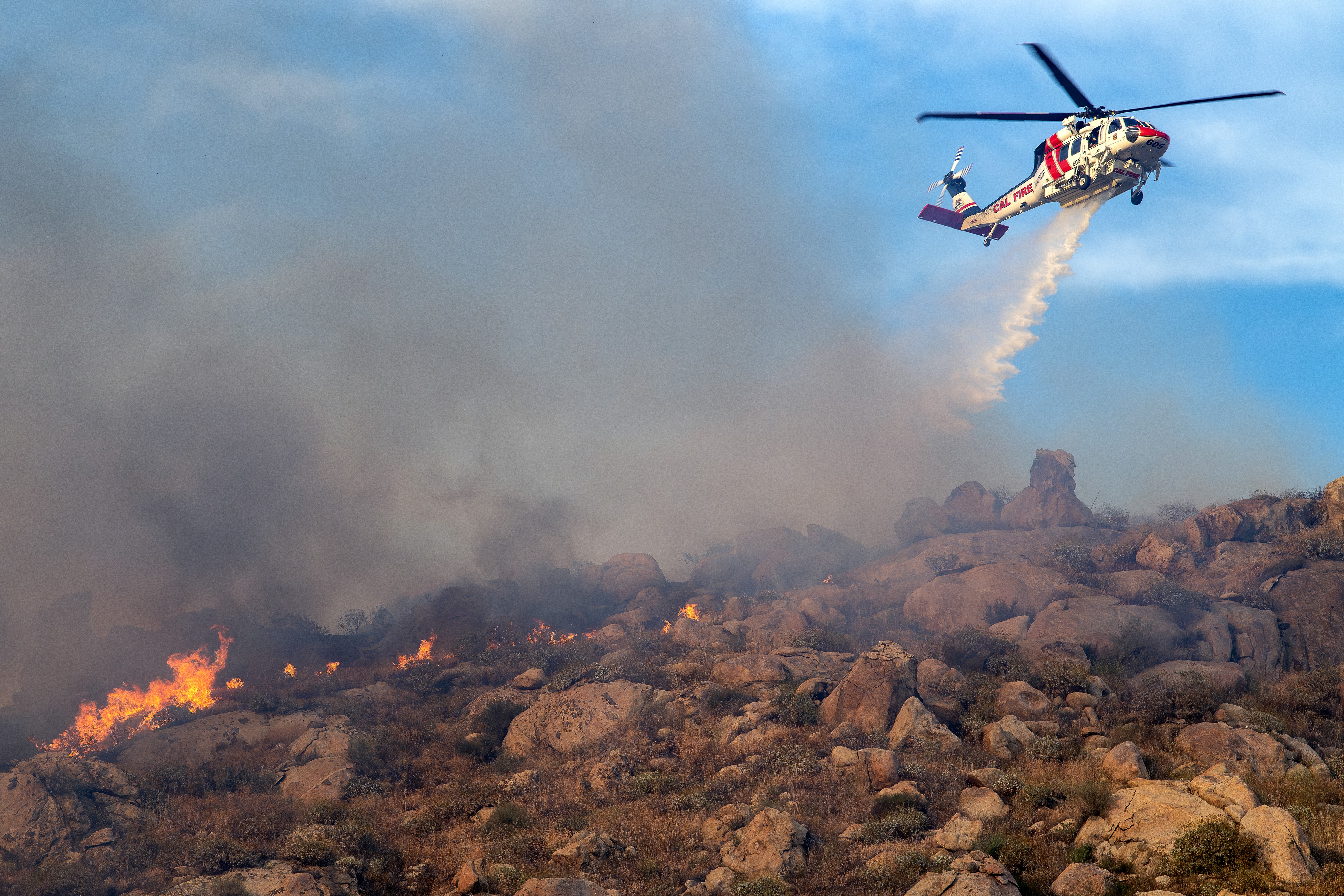 Cal Fire’s Firehawk drops on the Springs Fire in Lake Perris, California. The late afternoon light was very soft and perfectly illuminated the Firehawk and falling water against a beautiful blue sky.  I shot this photograph at a wider angle to include the smoke and burning landscape. I went on a short dirt road to get next to the fire and where the helicopters were dropping. I found the correct position using the Watch Duty app.