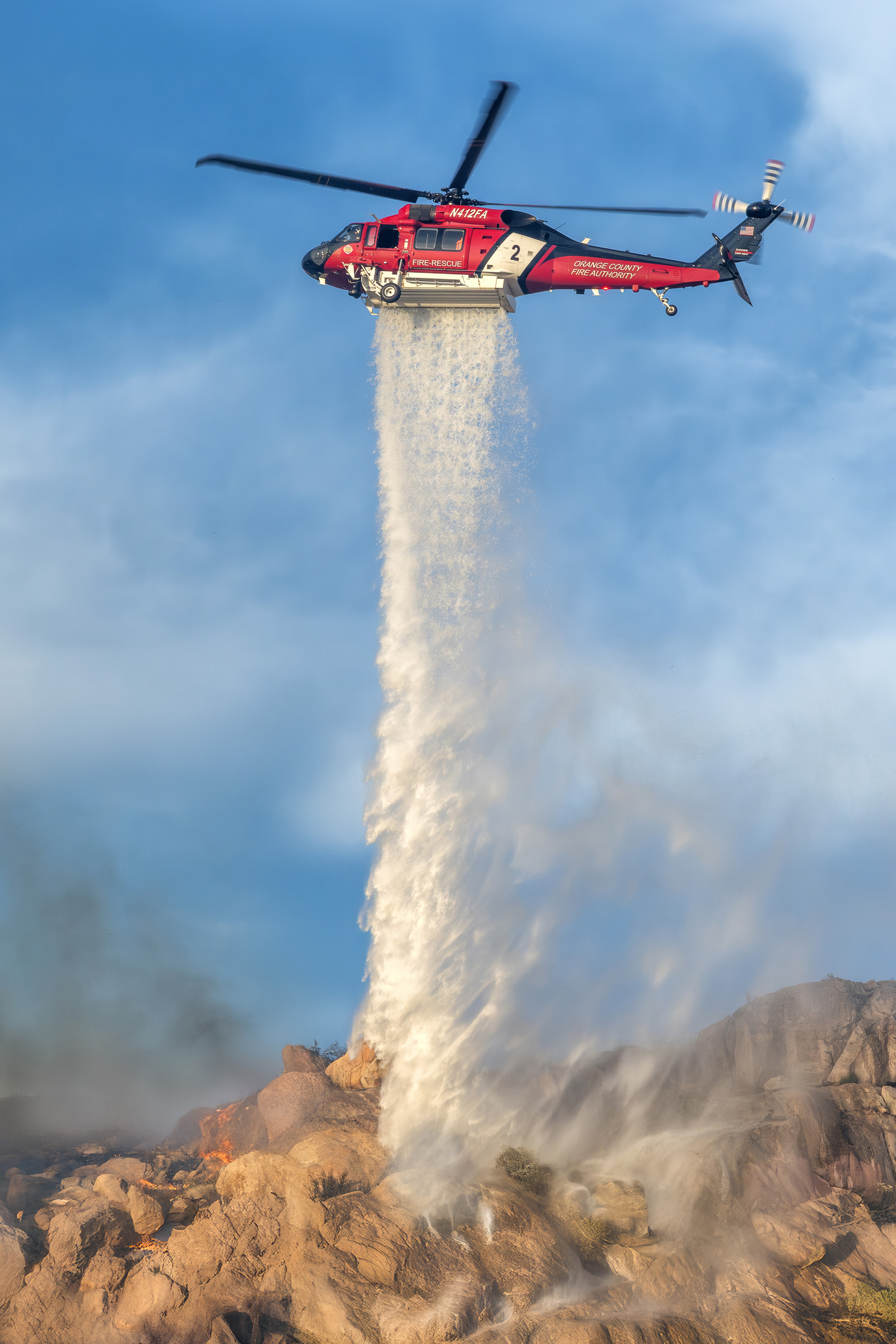 Orange County Fire Authority's Firehawk drops on the Springs Fire in Lake Perris, California. The late afternoon light was very soft and perfectly illuminated the Firehawk and falling water against a beautiful blue sky. The misting of the water at the bottom of photograph looks like its cascading like a waterfall.