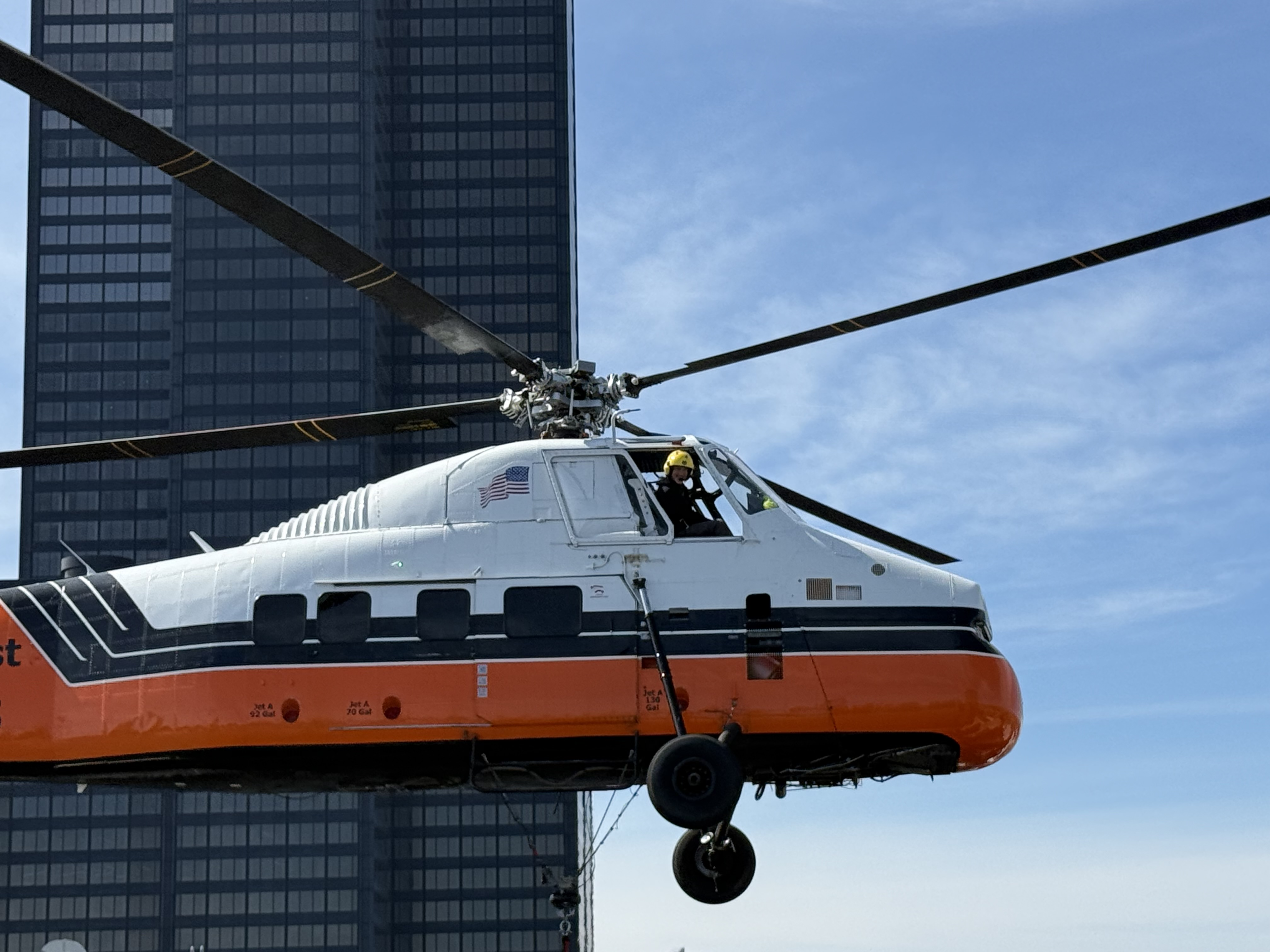 A Midwest Helicopter Airways Sikorsky S-58T works among the skyscrapers of downtown Chicago, setting rooftop equipment where traditional cranes can’t easily reach. Captured by the signalman from the rooftop, this close-up catches the pilot at work as the helicopter descends to pick up the next unit. The image offers a unique perspective of the precision, communication, and teamwork required between the pilot and ground crew during aerial lift operations in the heart of the city.