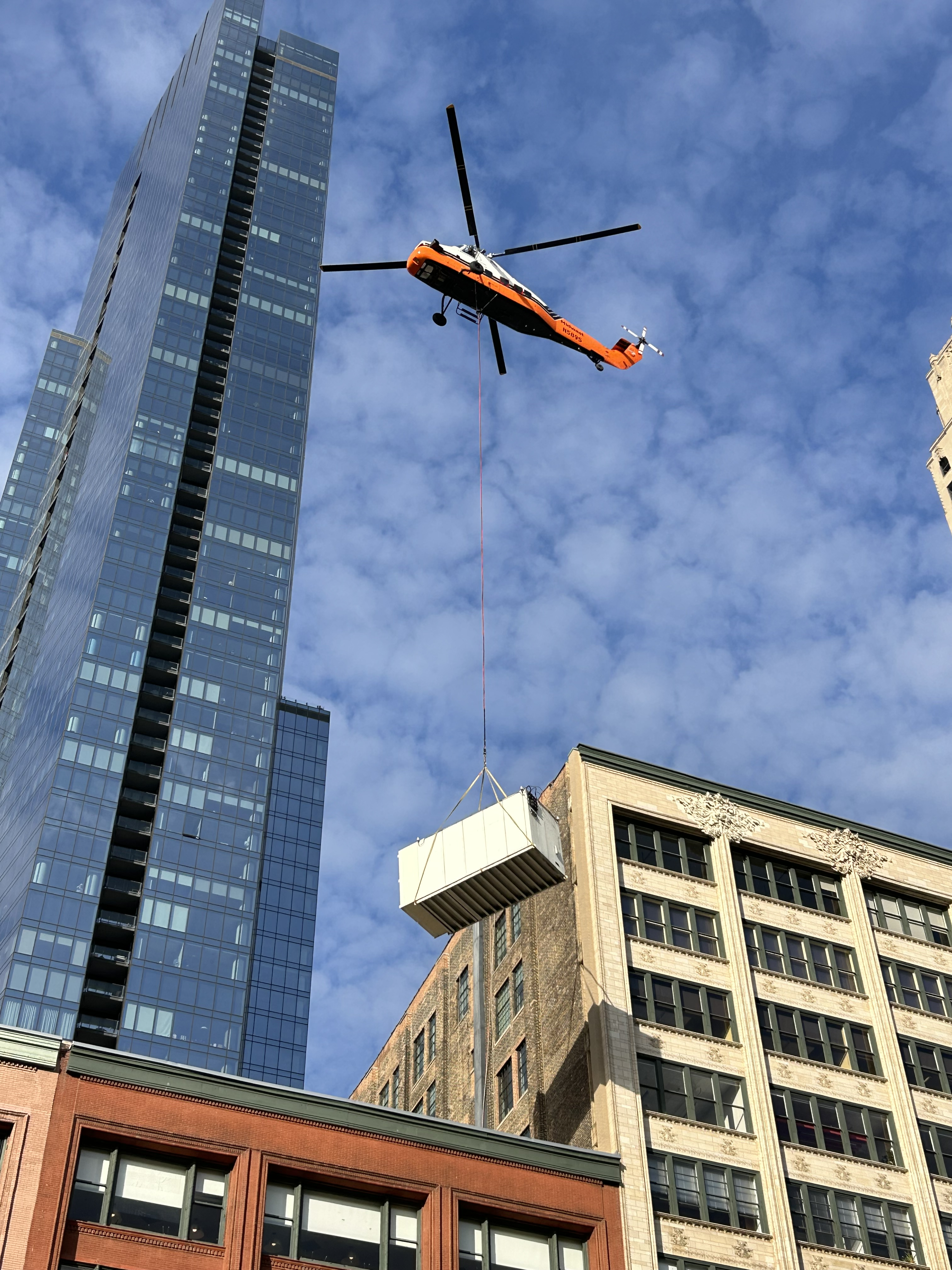 Midwest Helicopter Airways takes heavy lifting to new heights as the Sikorsky S-58T carries a large rooftop unit through the downtown Chicago skyline. Suspended beneath the helicopter on a longline, the unit is maneuvered above the surrounding buildings for precise placement on the rooftop. This photo captures the unique capability of aerial crane operations—moving large equipment efficiently in tight urban spaces where access from the ground can be a challenge.