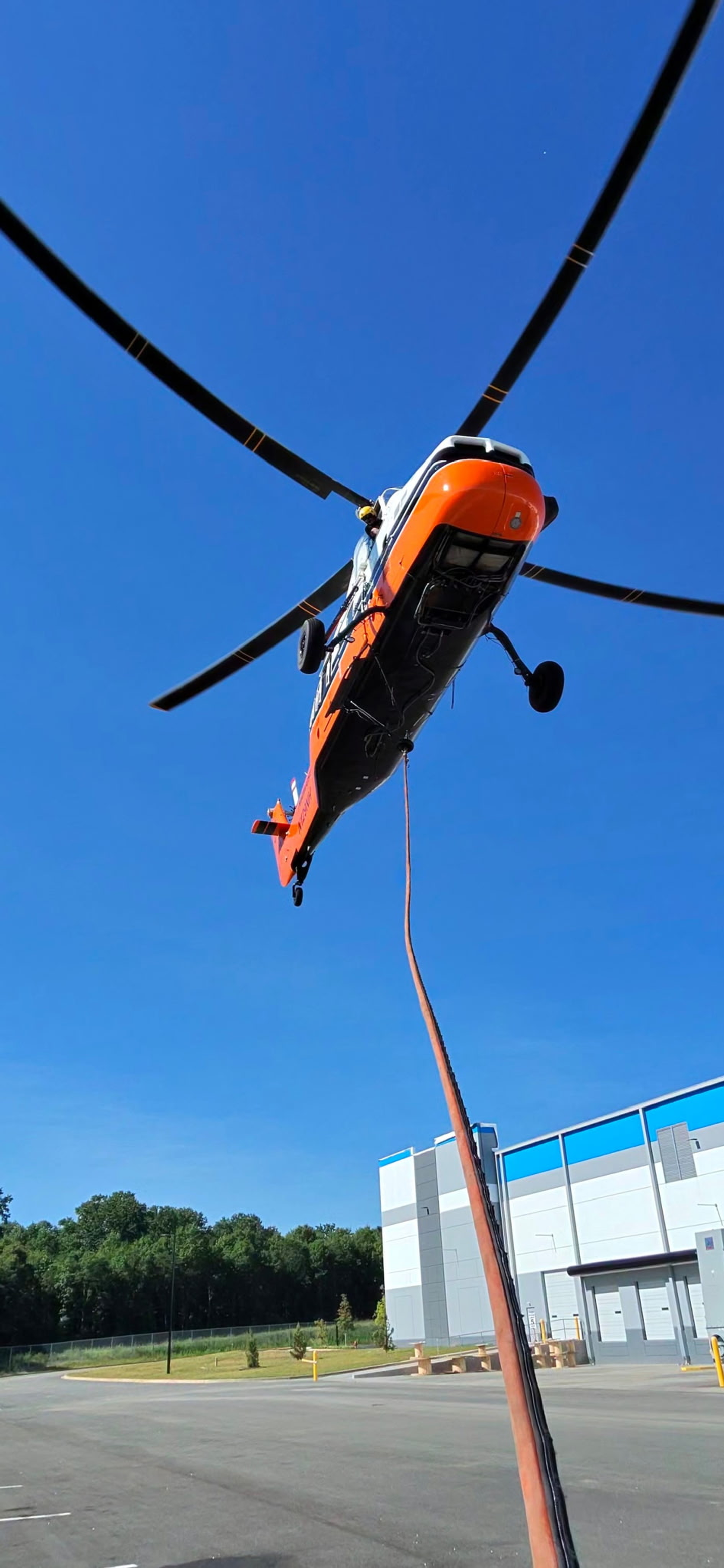 Midwest Helicopter Airways’ Sikorsky S-58T hovers overhead during an aerial lift operation, captured from directly beneath the aircraft. The longline stretches from the helicopter toward the staging area below as the pilot prepares for the next lift. This unique perspective showcases the size of the S-58T, the precision required to maneuver from above, and the specialized equipment that makes moving heavy loads quickly and efficiently possible.