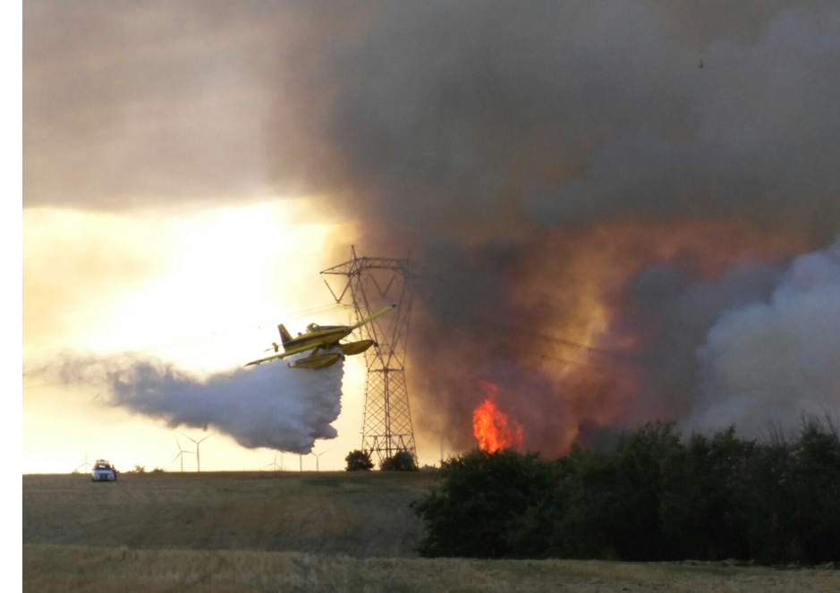 A yellow airplane is flying over a field with a fire in the background. AI generated content