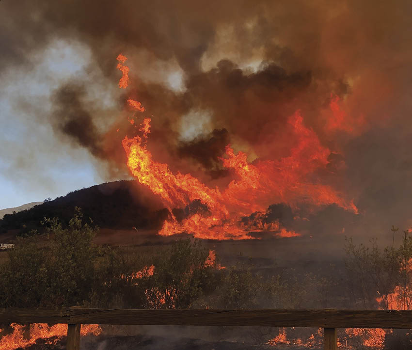 A large fire is burning in a field, with flames reaching high into the sky. The fire is surrounded by a fence, and there is a truck visible in the distance. AI generated content