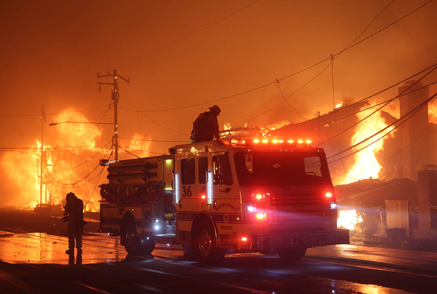 A fire truck is parked on a street with a fire burning in the background. AI generated content