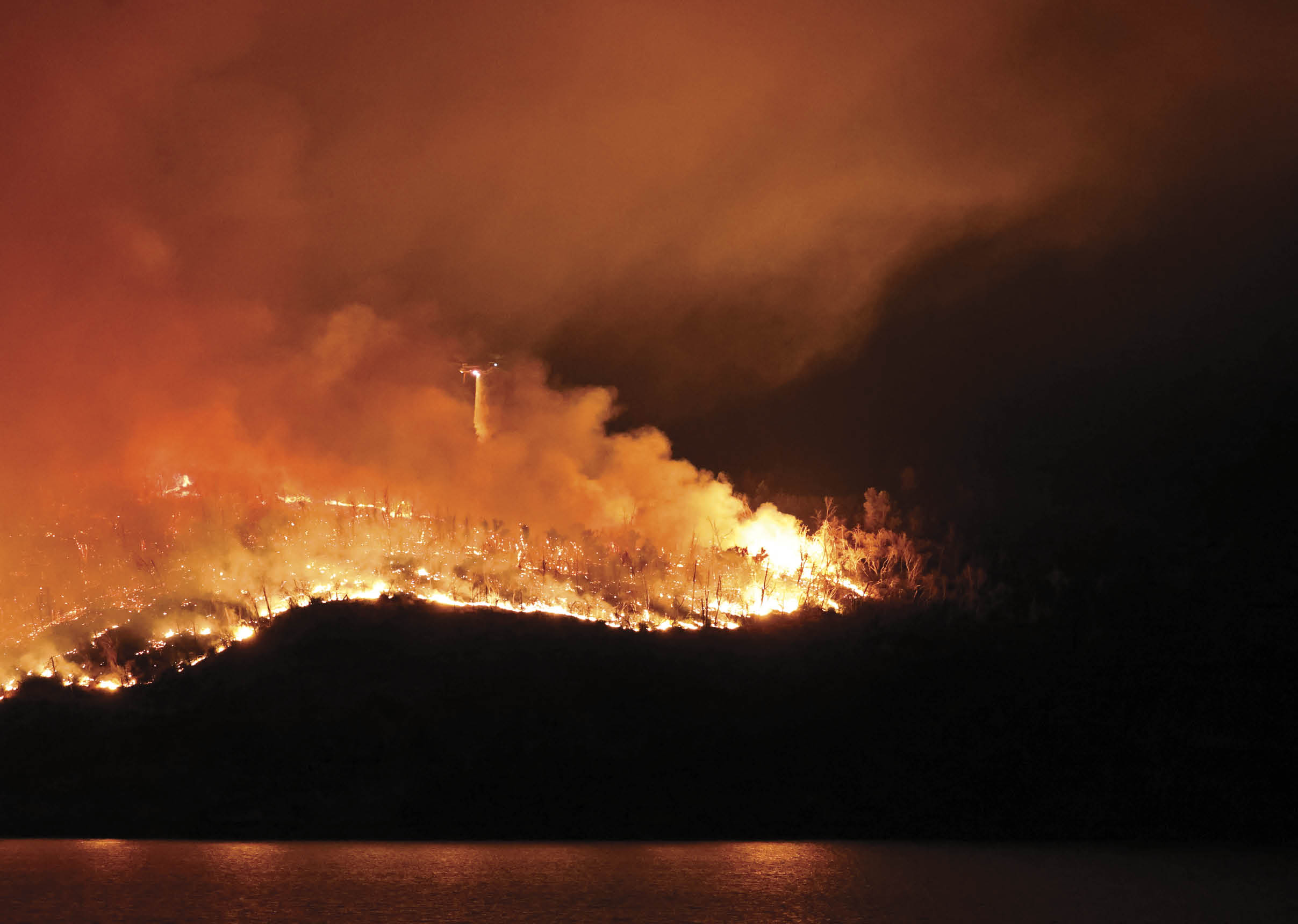 A large fire is burning in the distance, with flames reaching high into the sky. The fire is located near a body of water, creating a dramatic scene. AI generated content