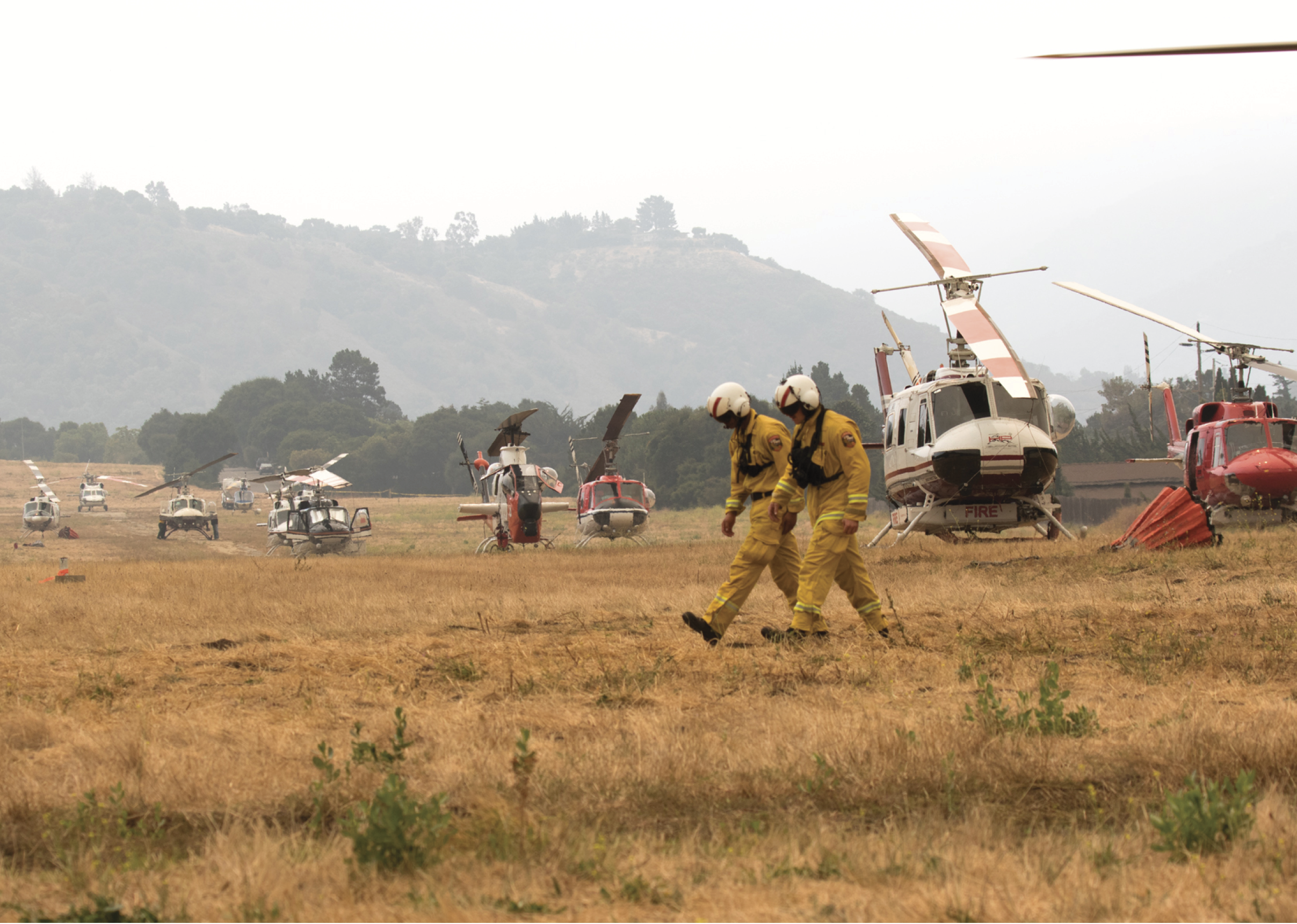 A man in a yellow jumpsuit is walking through a field with several helicopters in the background. AI generated content