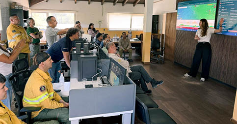 A group of people are gathered in a room, watching a large screen. There are several chairs, a dining table, and a couch in the room. A woman is standing in front of the screen, possibly giving a presentation. The people in the room are engaged in the presentation, with some of them holding cell phones. AI generated content
