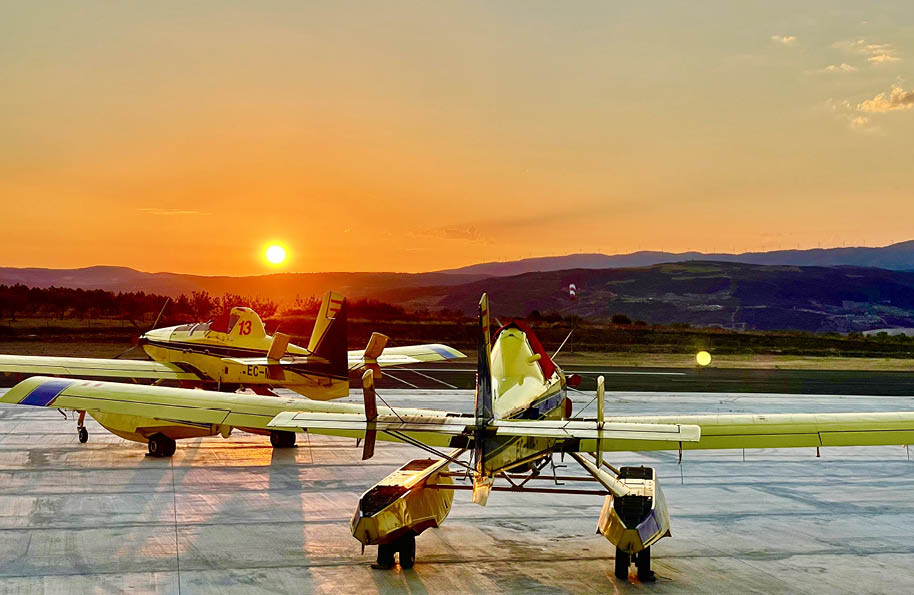 Two airplanes are parked on a runway, one is yellow and the other is white. The sun is setting in the background, casting a warm glow on the scene. AI generated content