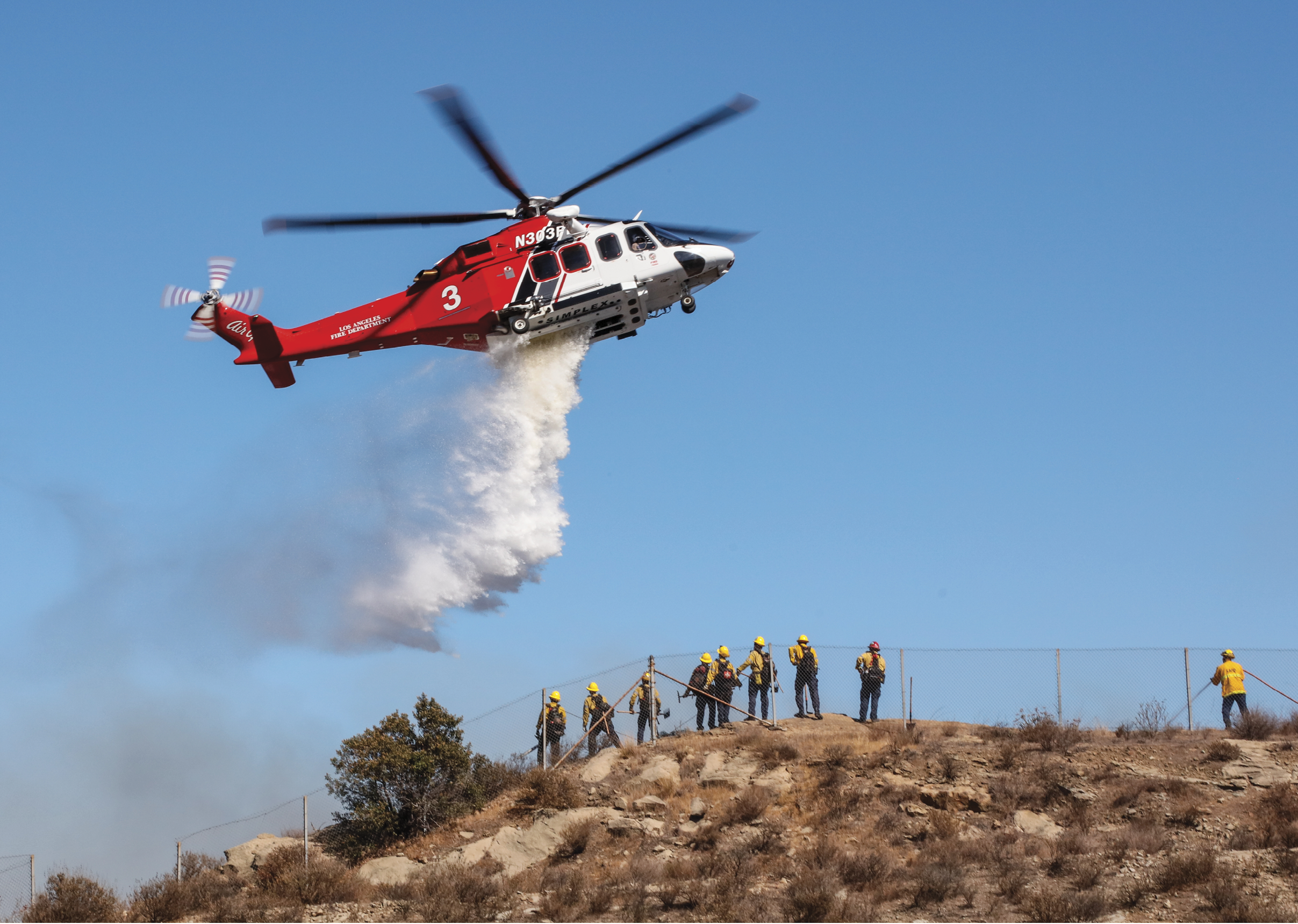 A helicopter is flying over a group of firefighters on a hill, spraying water to help them fight a fire. AI generated content