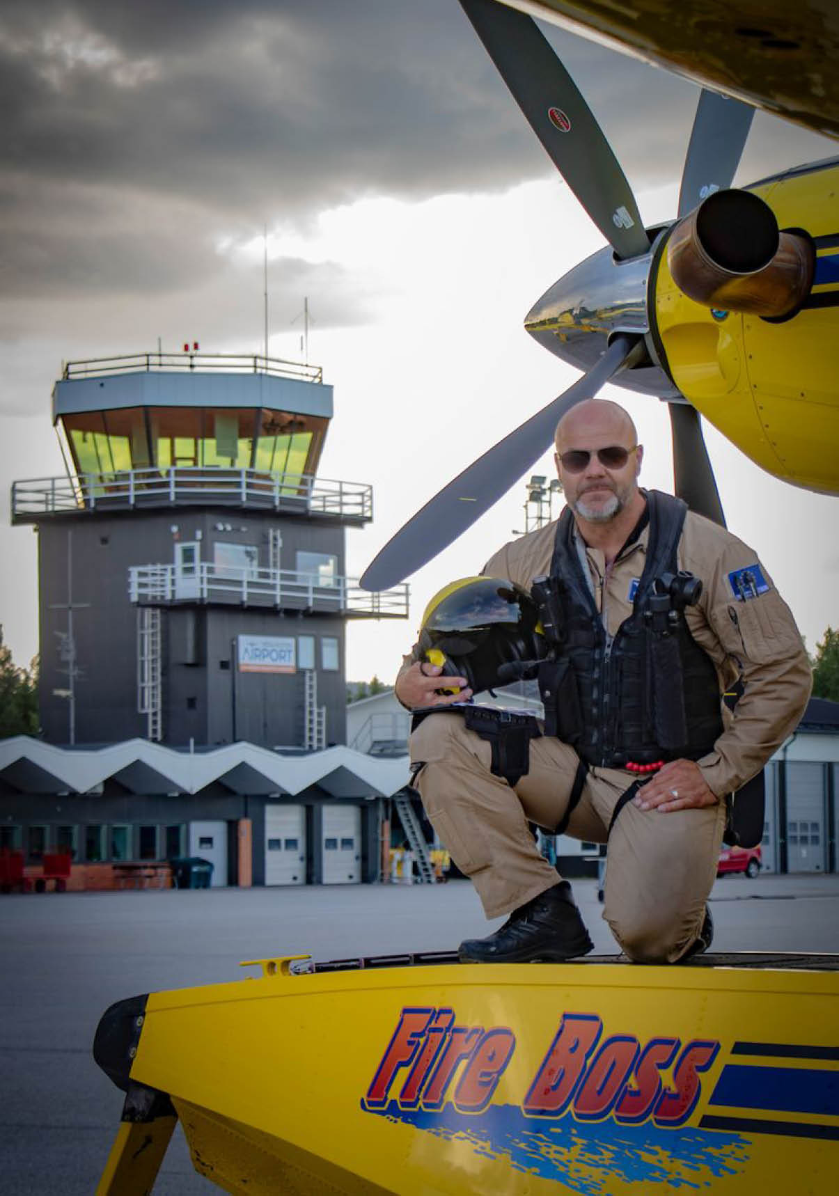 A person wearing a flight suit and sunglasses is sitting next to a propeller airplane with an airport control tower in the background under a cloudy sky. Description generated by AI