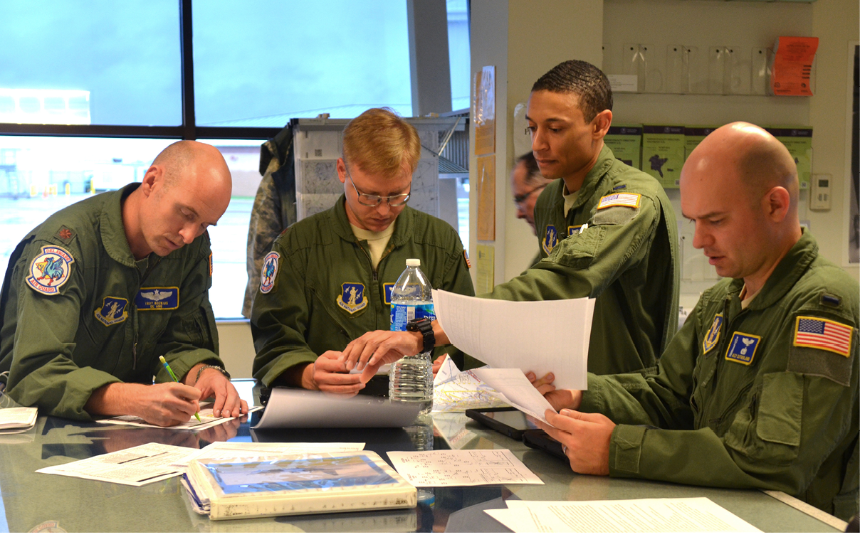 Two adults in military uniforms are seated at a table, one writing on paper and the other holding a water bottle while looking down. Description generated by AI