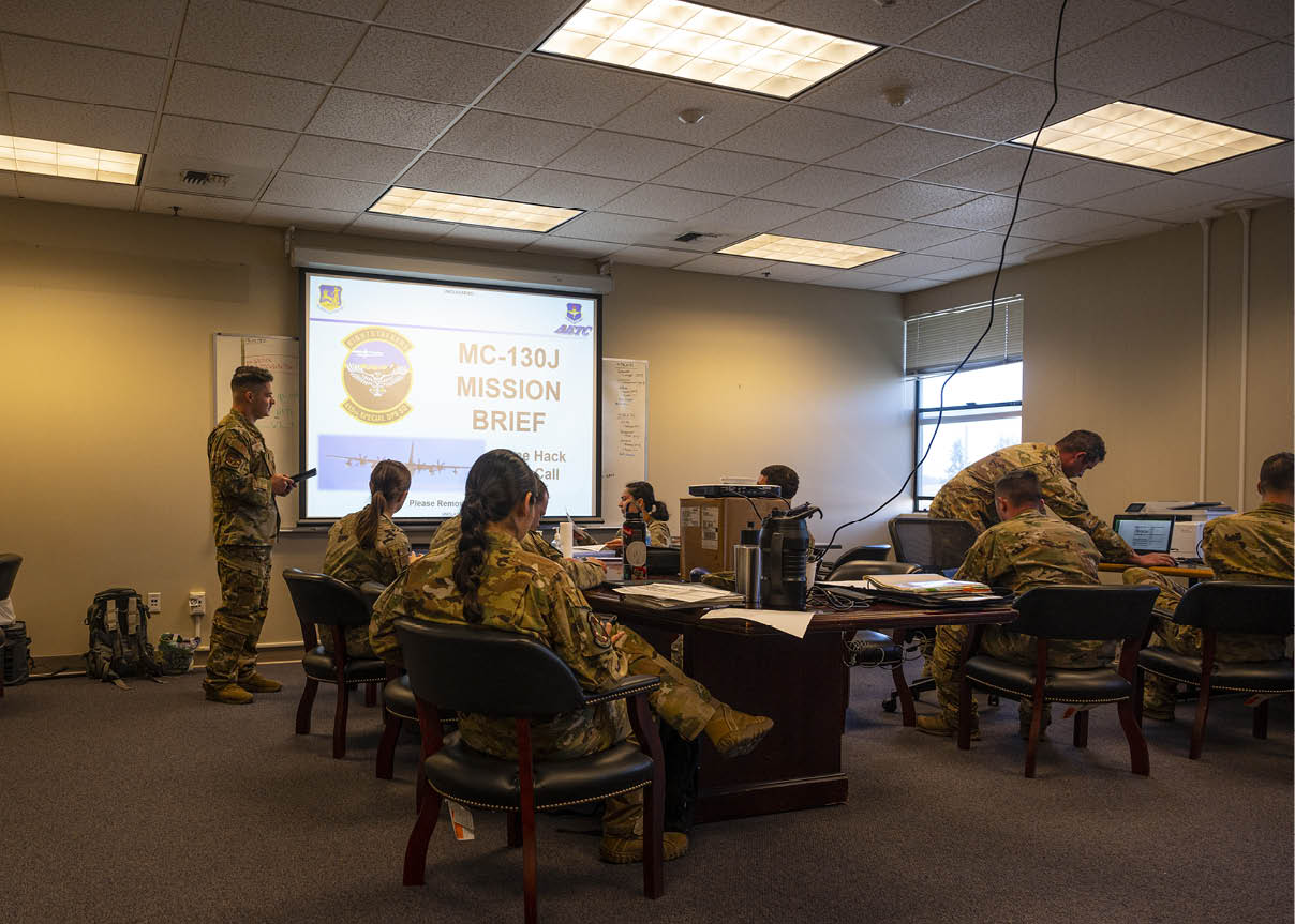 A person in military uniform is standing near a projection screen displaying an emblem and text in a room with a drop ceiling and fluorescent lighting. Description generated by AI