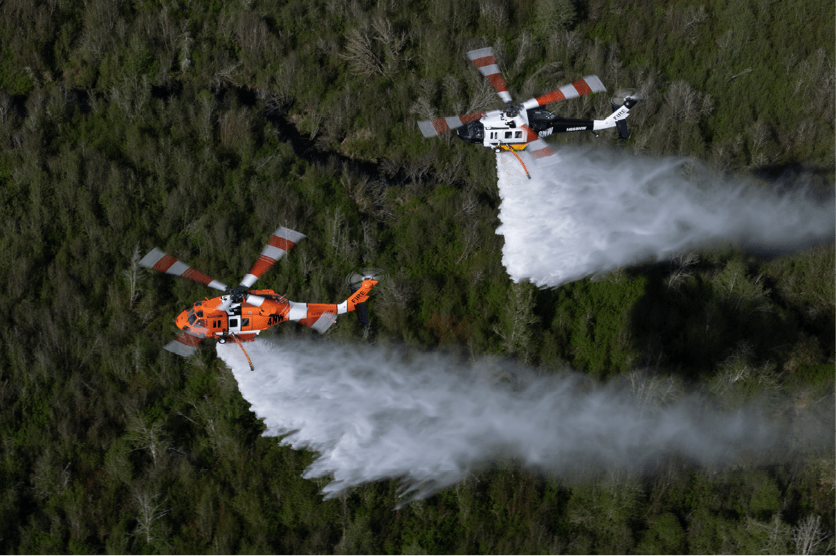 Two helicopters are flying over a forest while releasing large streams of water, likely to extinguish a wildfire. Description generated by AI
