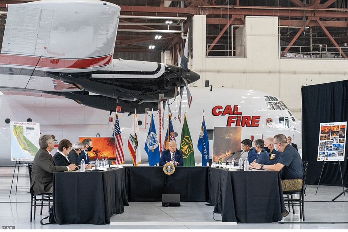 People seated at tables in a hangar are engaged in a discussion with a large airplane and CAL FIRE signage in the background. Description generated by AI