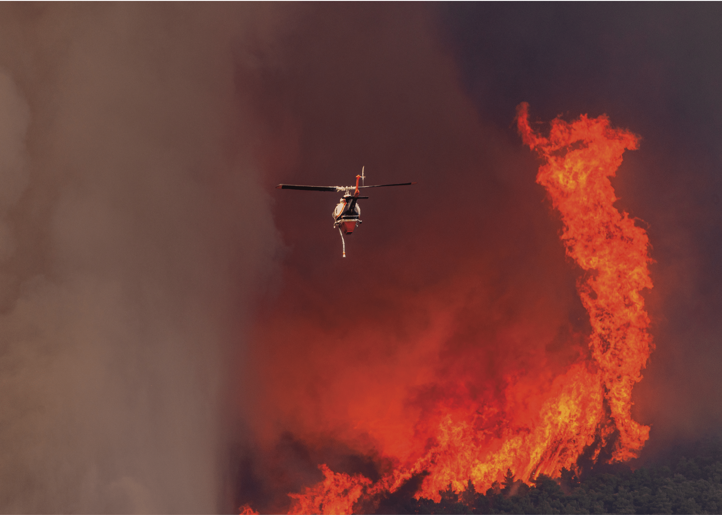 A helicopter is flying in front of a large, intense wildfire with thick smoke and bright orange flames. Description generated by AI