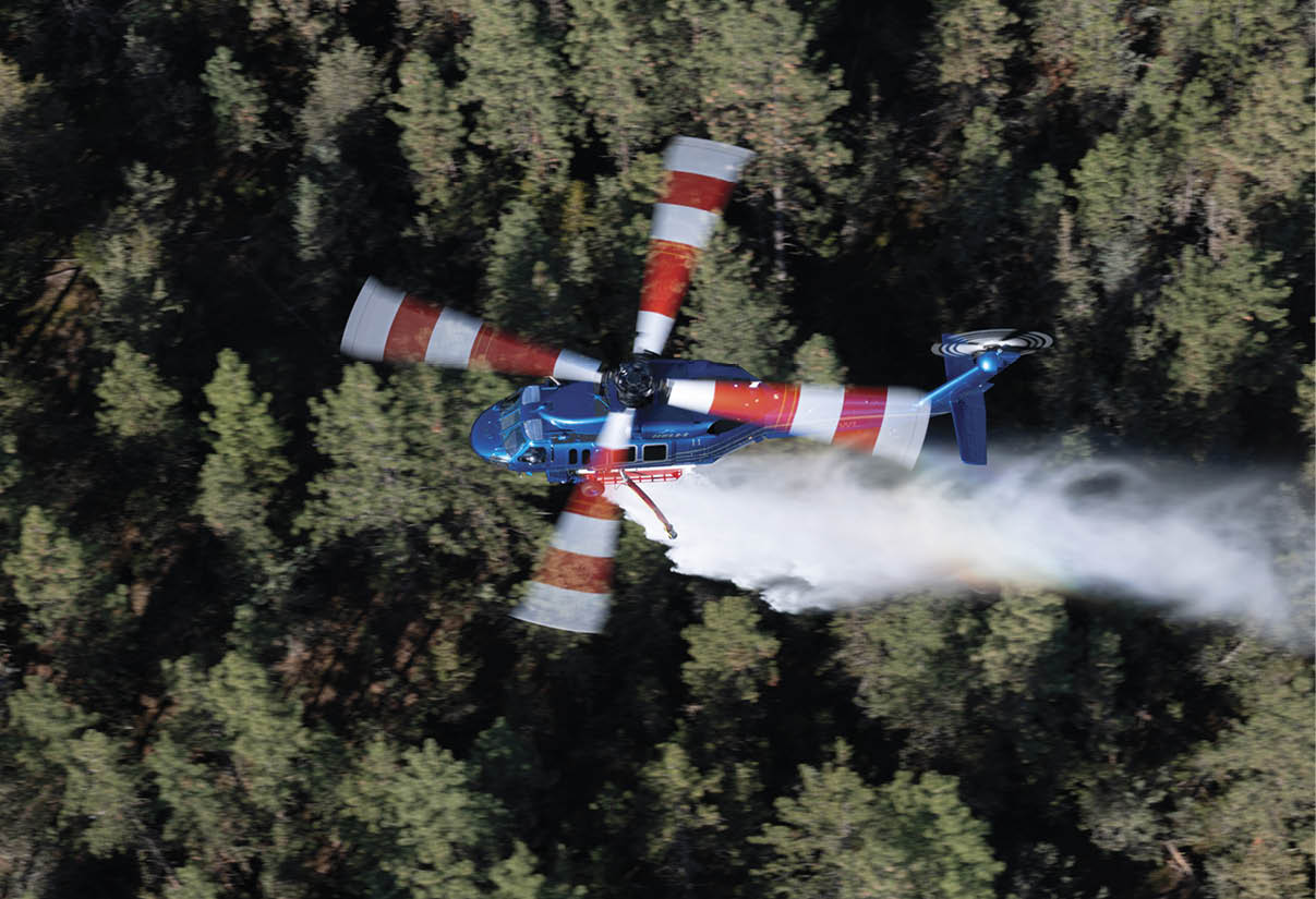 A helicopter with red and white striped rotor blades is flying upside down above a forest, emitting a trail of white smoke. Description generated by AI