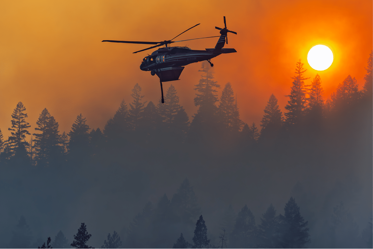A helicopter flies over a forest with smoke and an orange sky as the sun sets or rises in the background. Description generated by AI