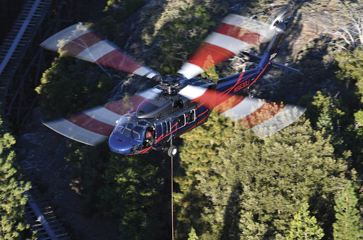 A helicopter with spinning rotor blades is flying above a forested area with a cable hanging below it. Description generated by AI