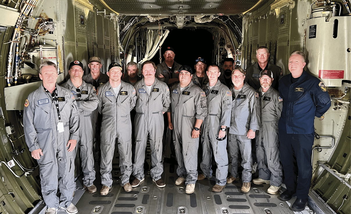 A group of people wearing gray flight suits and one person in a dark blue uniform stand inside the cargo area of an aircraft with exposed wiring and structural elements. Description generated by AI