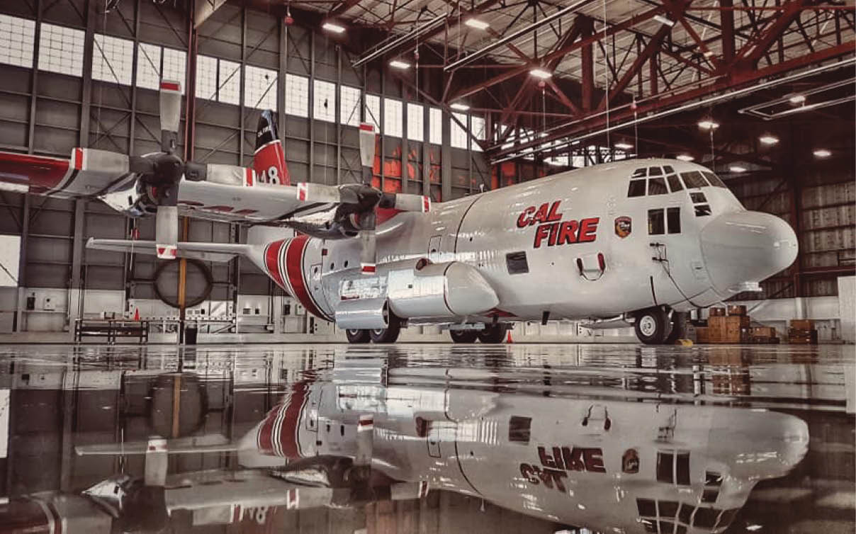 A large CAL FIRE aircraft is parked inside a spacious hangar with its reflection visible on the shiny floor. Description generated by AI