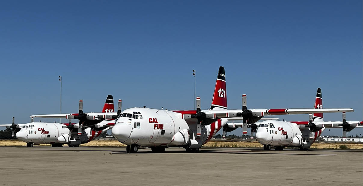 Three CAL FIRE aircraft with red and white markings are parked on a tarmac under a clear blue sky. Description generated by AI