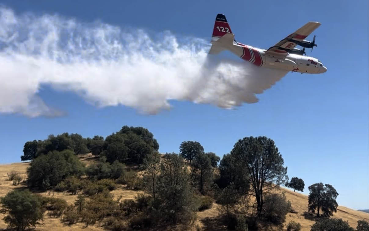 A firefighting airplane is releasing a large plume of water or fire retardant over a hilly, tree-covered landscape under a clear blue sky. Description generated by AI