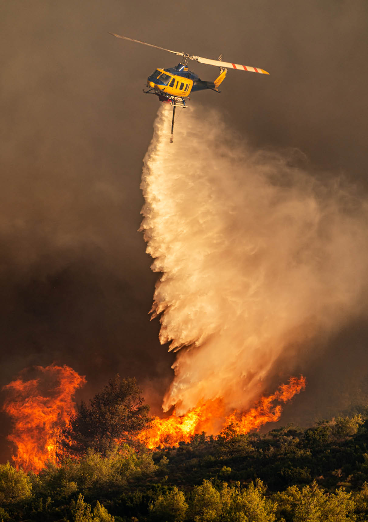 A helicopter is releasing water over a forest fire with large flames and smoke rising from the trees. Description generated by AI