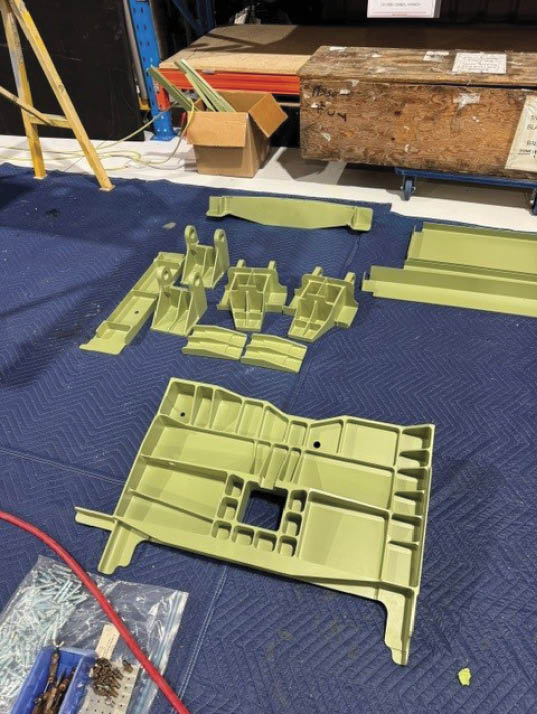 Several green plastic parts are arranged on a blue padded surface in a workshop setting with a wooden crate and cardboard box in the background. Description generated by AI