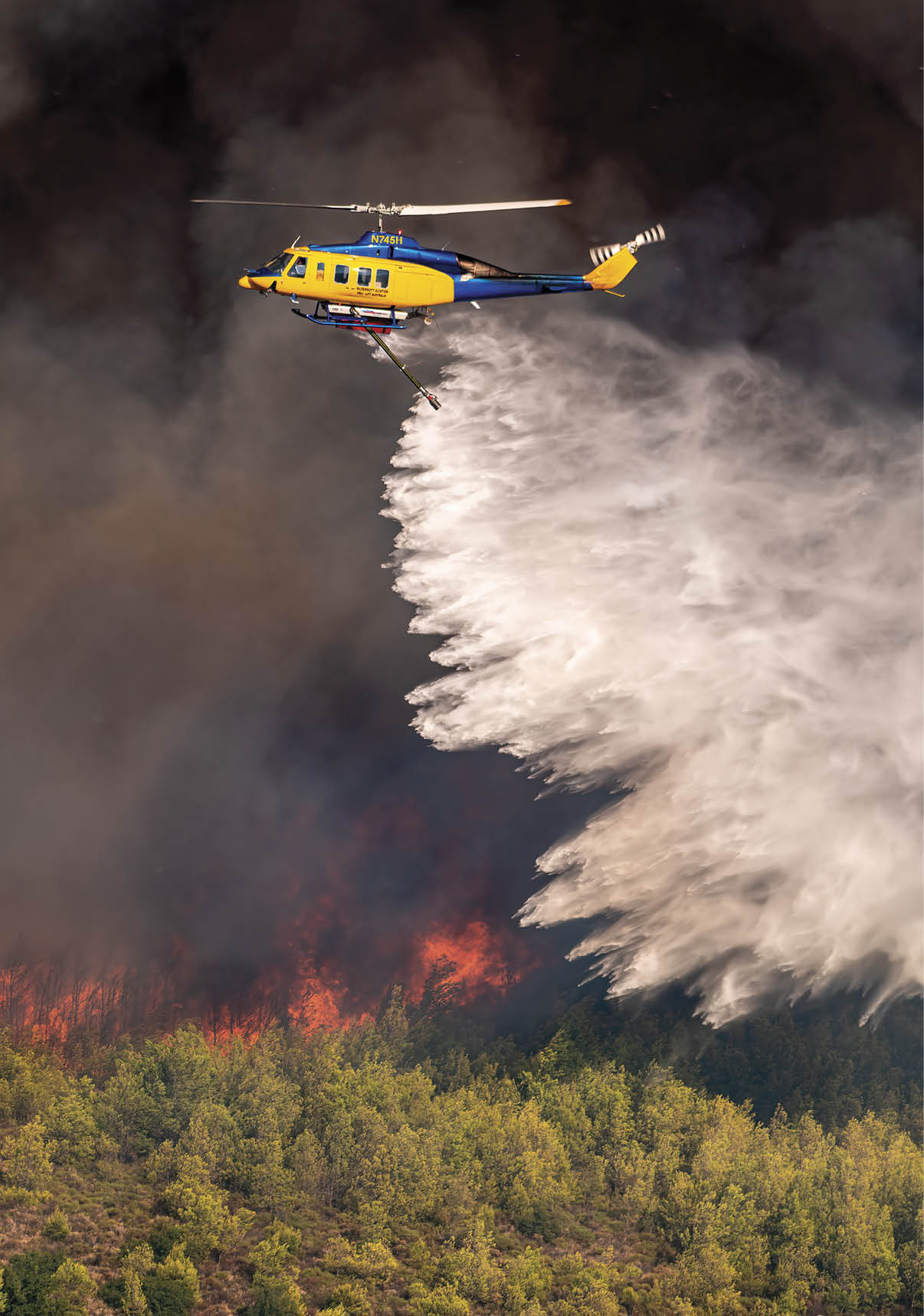 A helicopter is releasing a large spray of water over a forest fire with thick smoke and visible flames. Description generated by AI