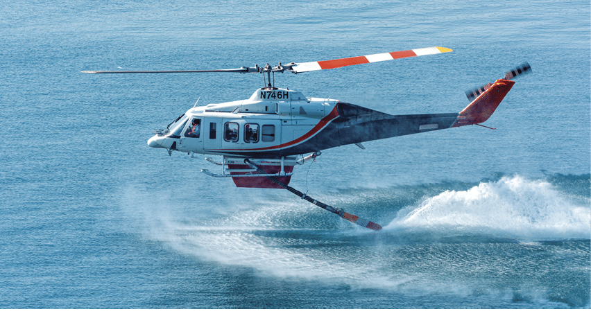 A helicopter with red and white rotor blades is flying low over water, creating splashes beneath it. Description generated by AI