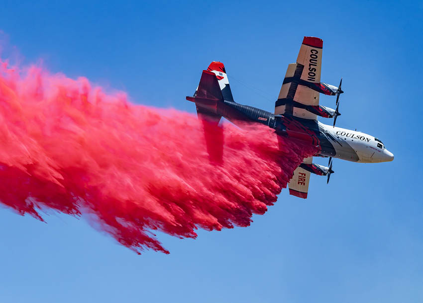 A firefighting airplane releases a large plume of red fire retardant against a clear blue sky. Description generated by AI