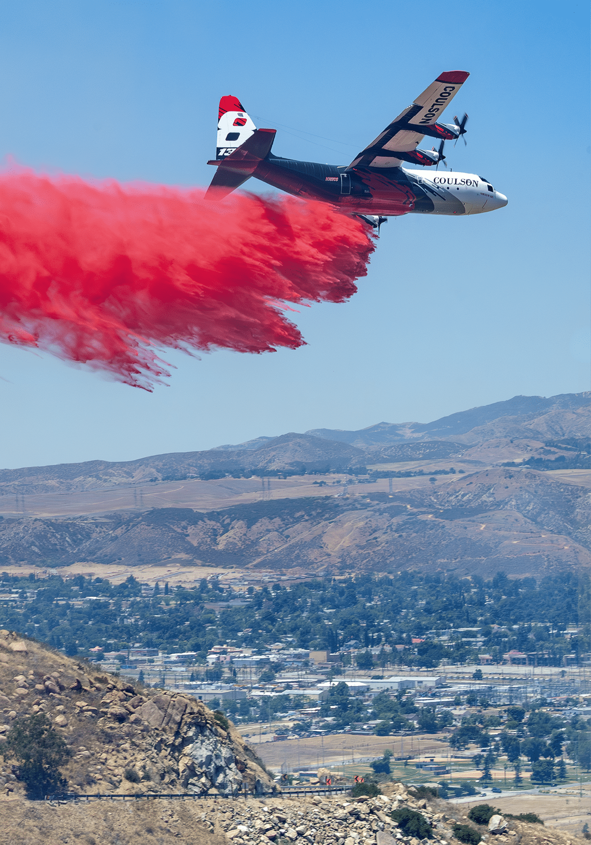 A firefighting airplane is releasing a large plume of red fire retardant over a mountainous area near a city. Description generated by AI