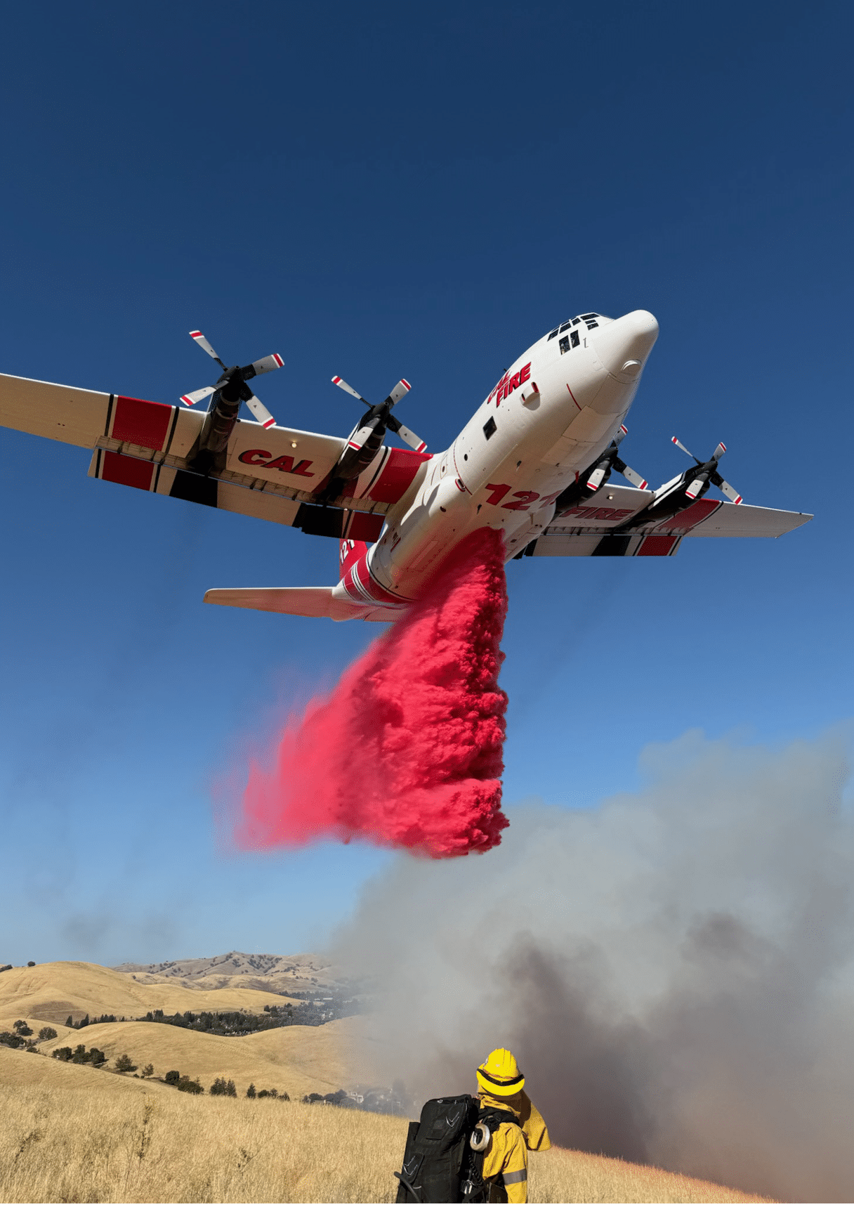 A firefighting airplane is releasing red fire retardant over a dry grassy area while a person in yellow protective gear watches from below near smoke. Description generated by AI
