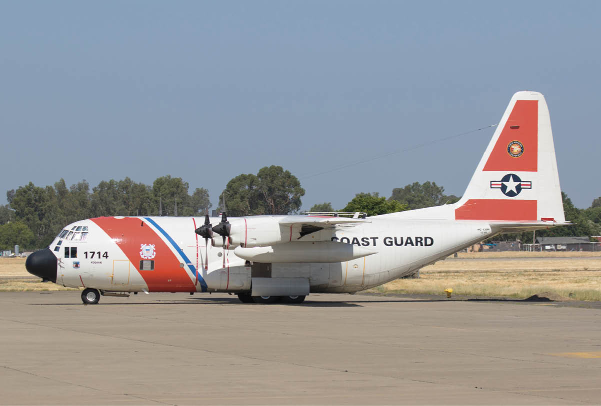 A Coast Guard airplane with red and white markings is parked on a runway with trees in the background. Description generated by AI