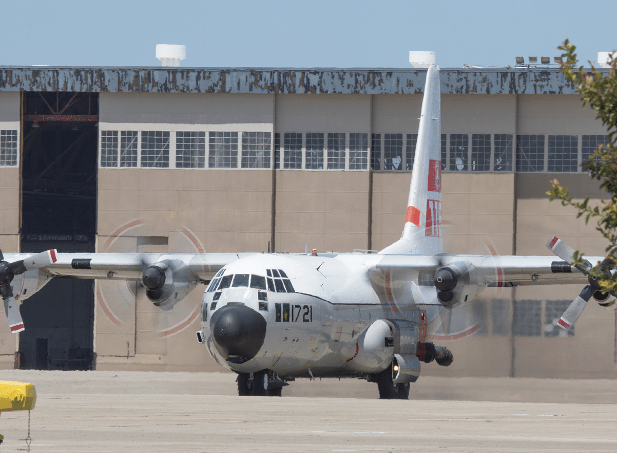 A military transport aircraft with spinning propellers is positioned on a runway in front of a large hangar building. Description generated by AI