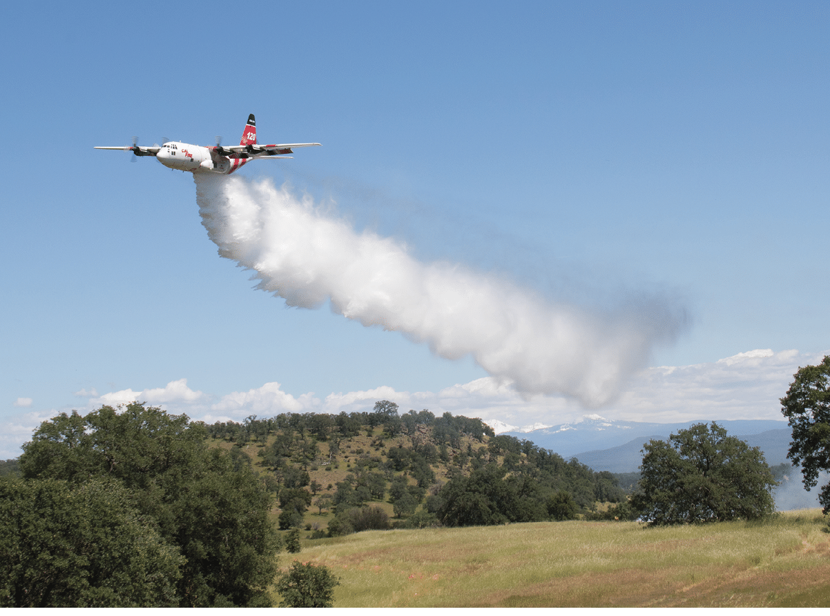 A firefighting airplane is releasing a large plume of water or fire retardant over a grassy and wooded landscape under a clear blue sky. Description generated by AI
