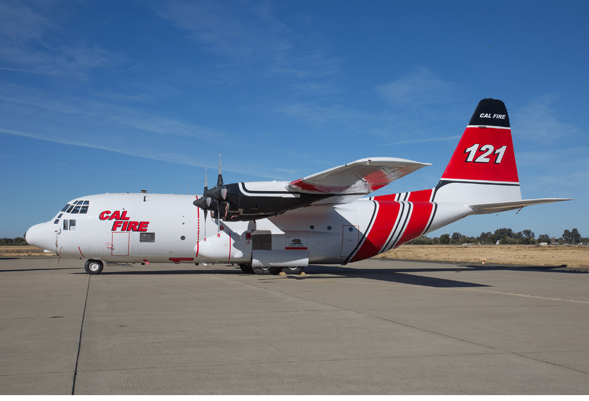A white and red CAL FIRE airplane with the number 121 is parked on a concrete runway under a clear blue sky. Description generated by AI