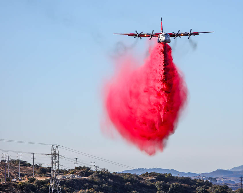 A firefighting airplane is releasing a large plume of red fire retardant over a mountainous area with power lines visible below. Description generated by AI