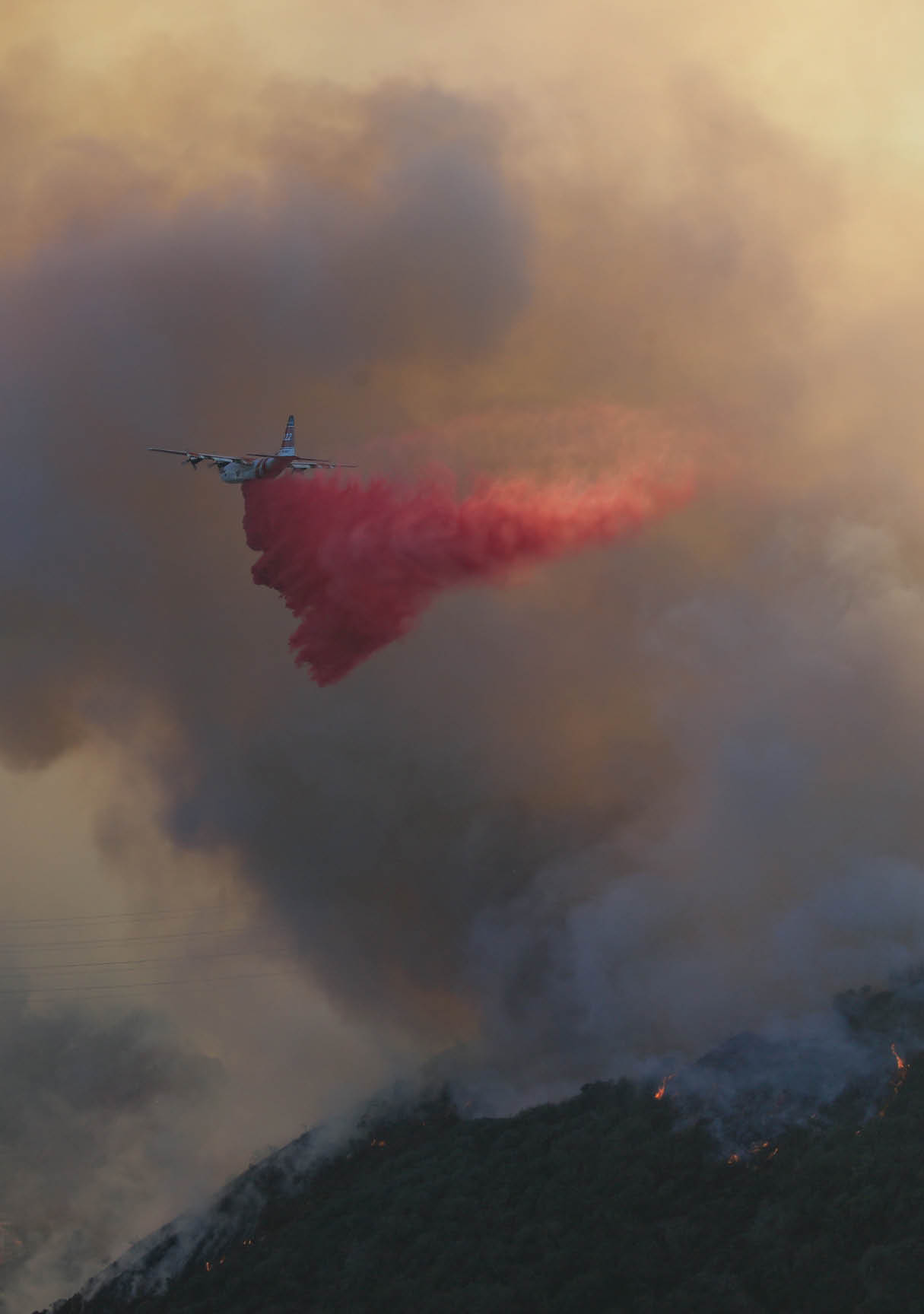 A plane is releasing a large plume of red smoke against a cloudy sky during what appears to be a firefighting or aerial display operation. Description generated by AI