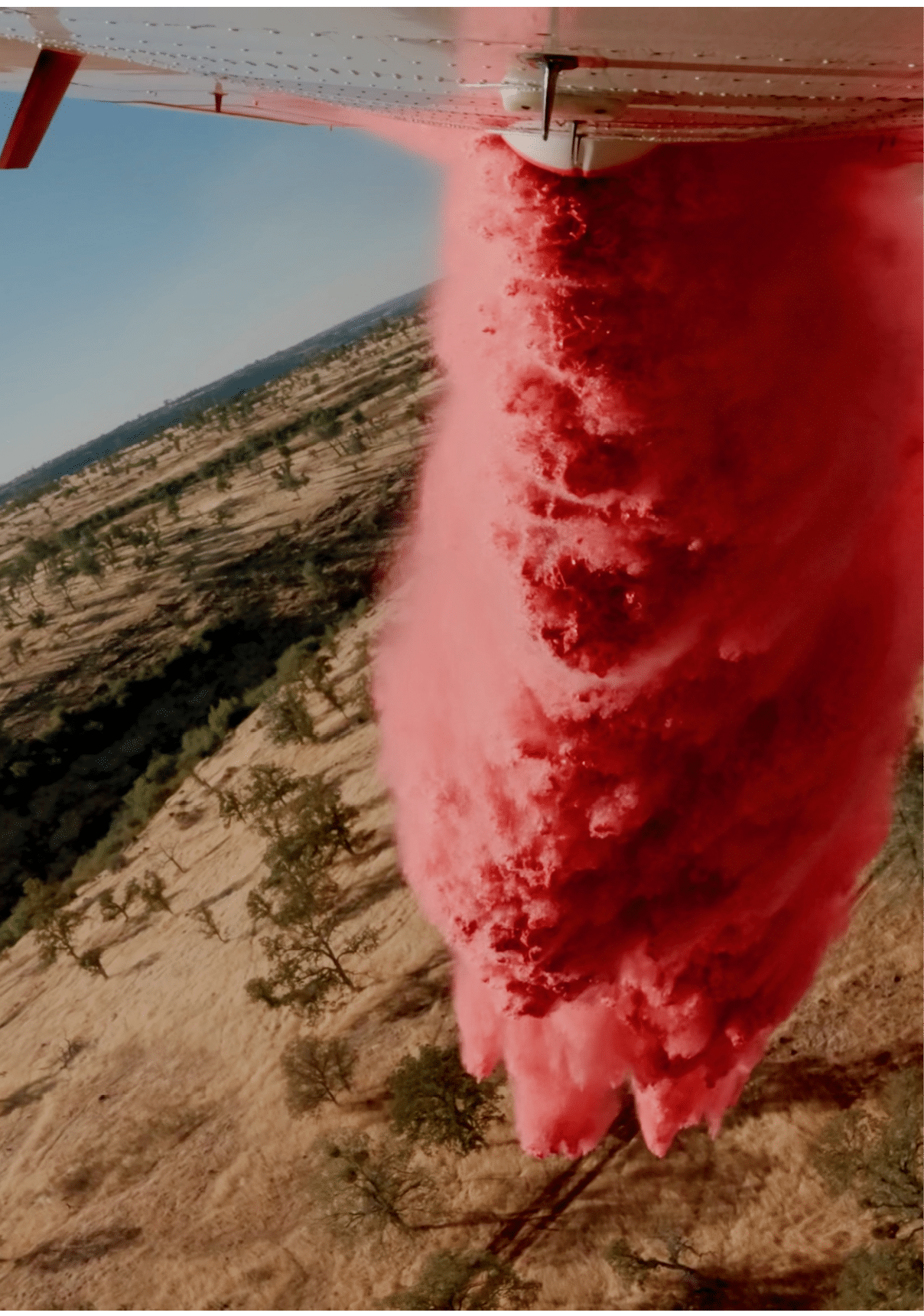 A large plume of red smoke or powder is rising vertically over a dry, sparsely vegetated landscape under a clear sky. Description generated by AI