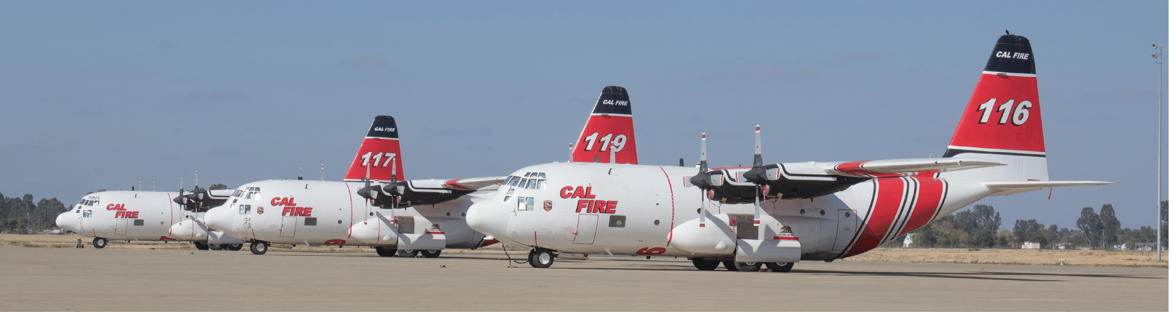 Four large white and red firefighting aircraft are lined up on a runway under a clear sky. Description generated by AI