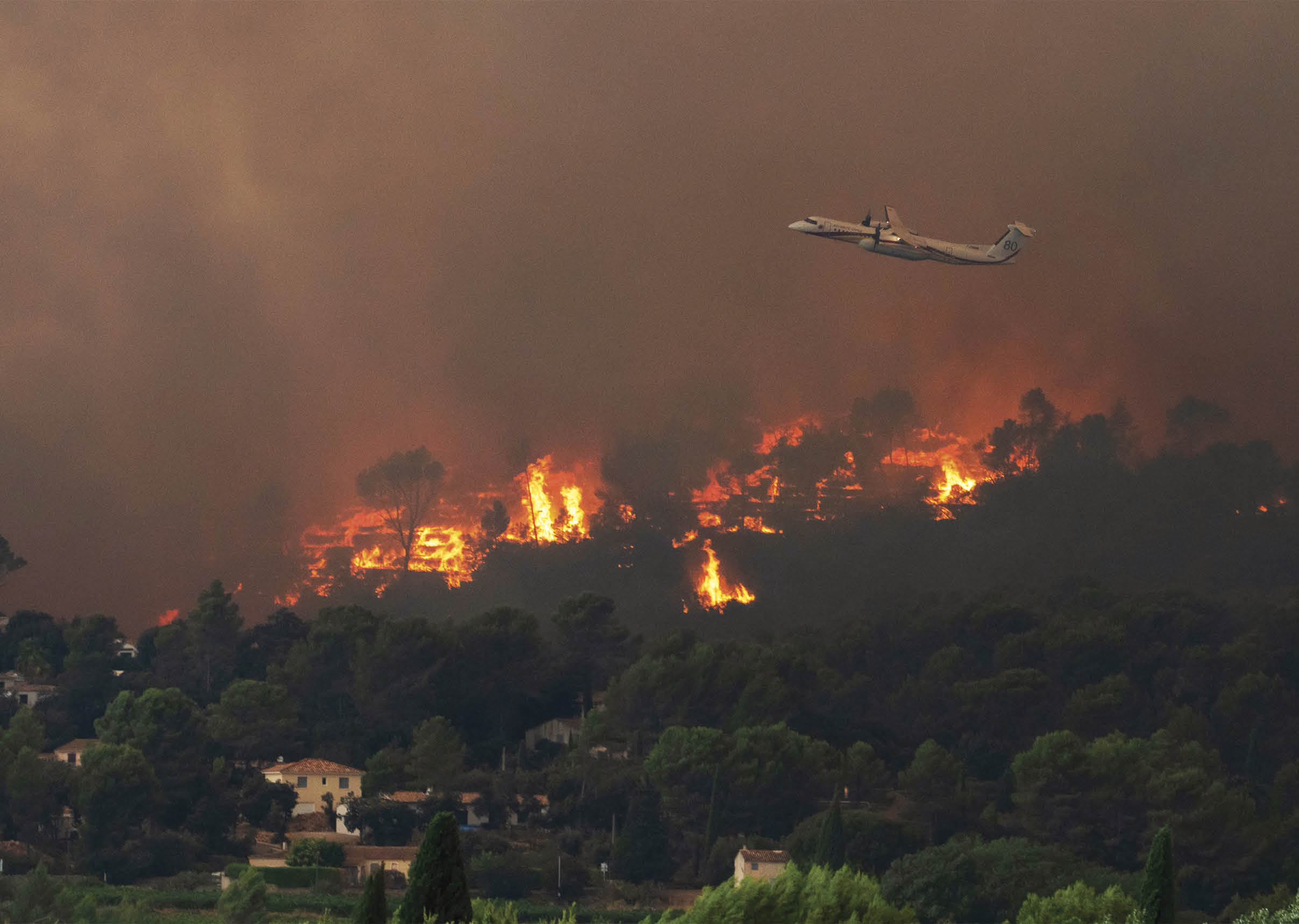 A wildfire burns on a hillside with thick smoke in the sky and a plane flying above the flames. Description generated by AI