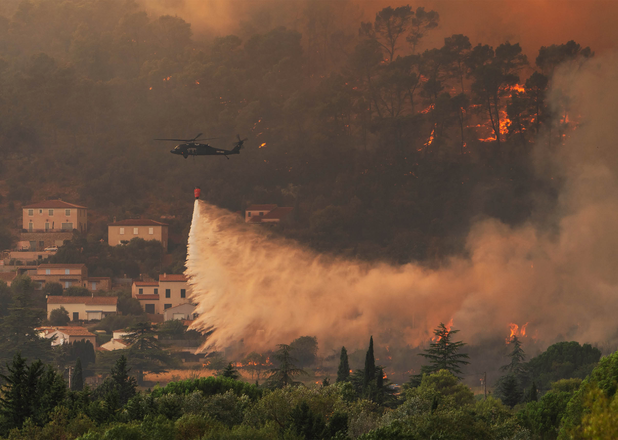 A helicopter is dropping water over a forest fire near a residential area with houses and trees. Description generated by AI