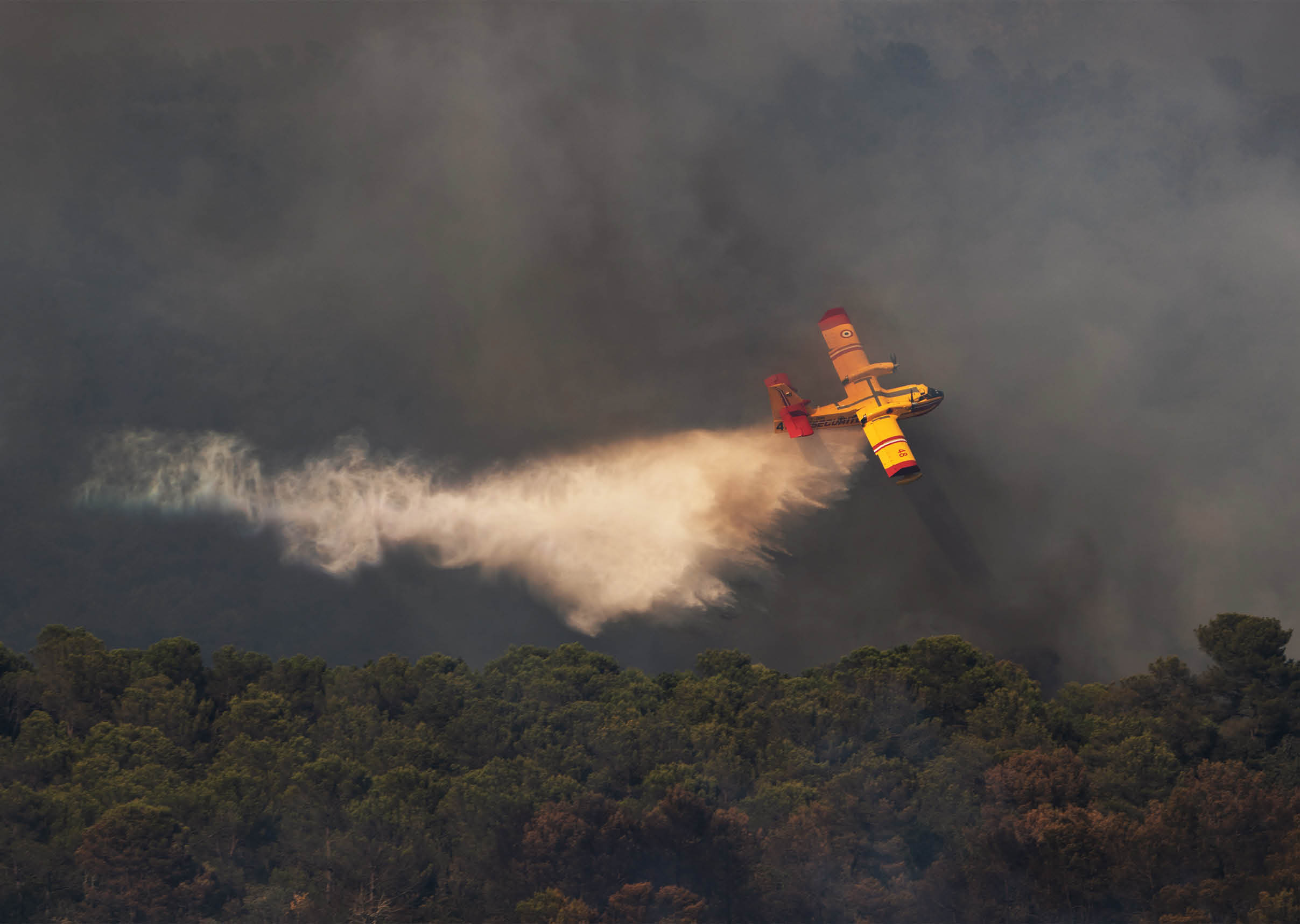 A yellow and red firefighting plane is releasing water over a forest fire with smoke rising in the background. Description generated by AI