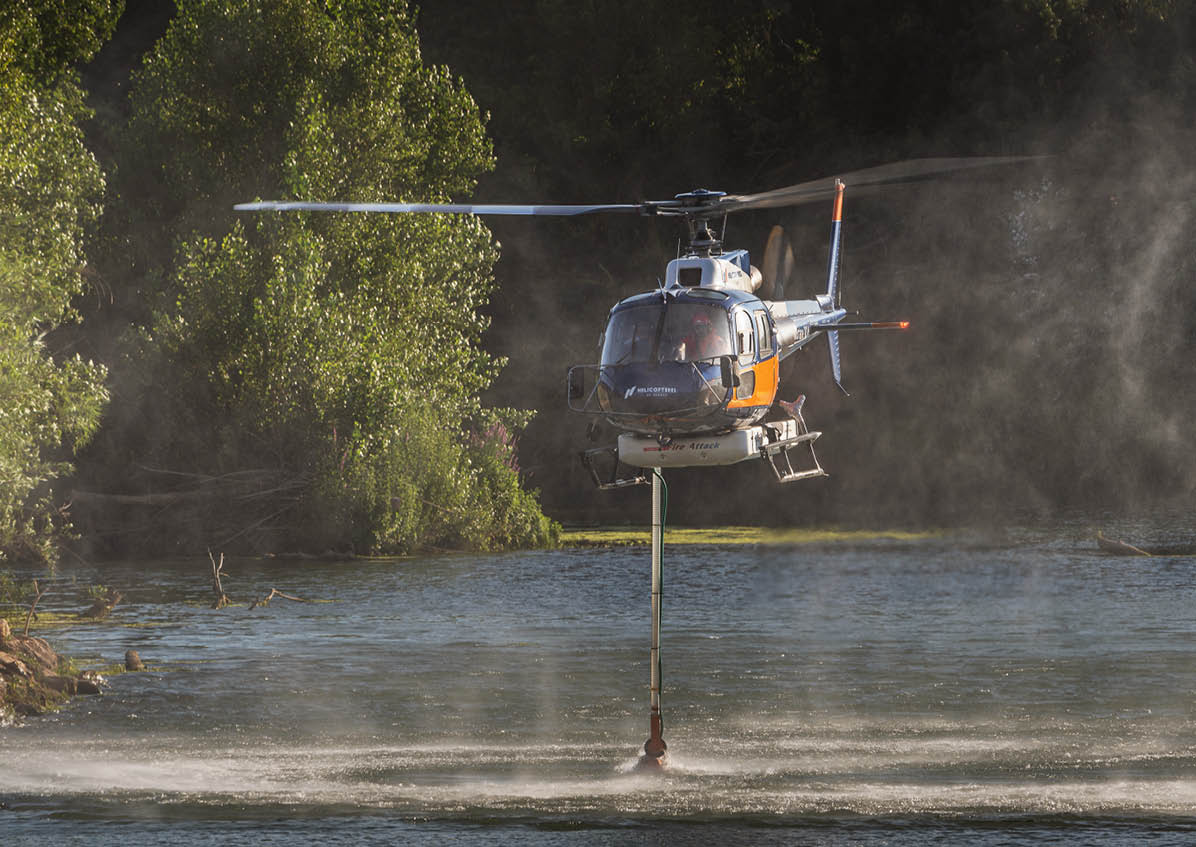 A helicopter is hovering above a body of water with a long hose or pipe extending down into the water, surrounded by trees and mist. Description generated by AI