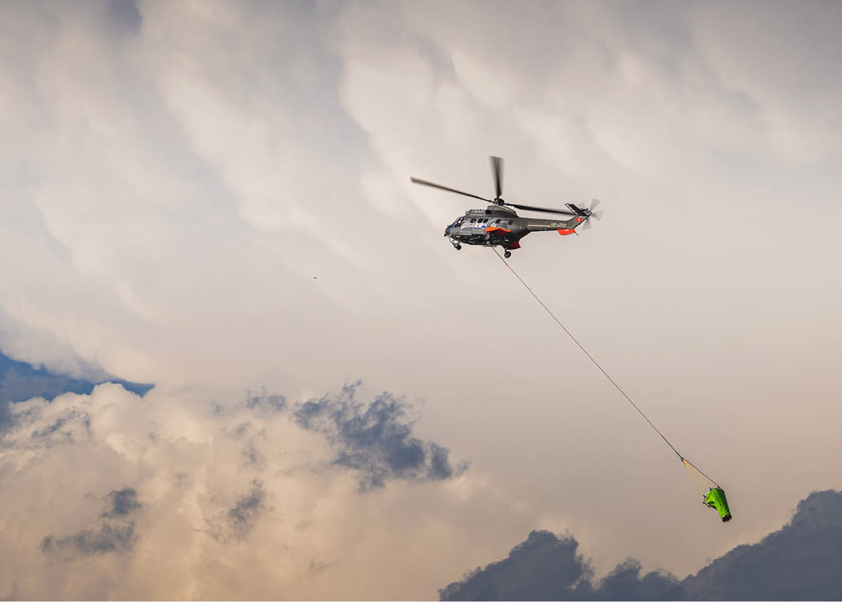A helicopter is flying in the sky carrying a green object suspended by a long cable against a backdrop of clouds. Description generated by AI